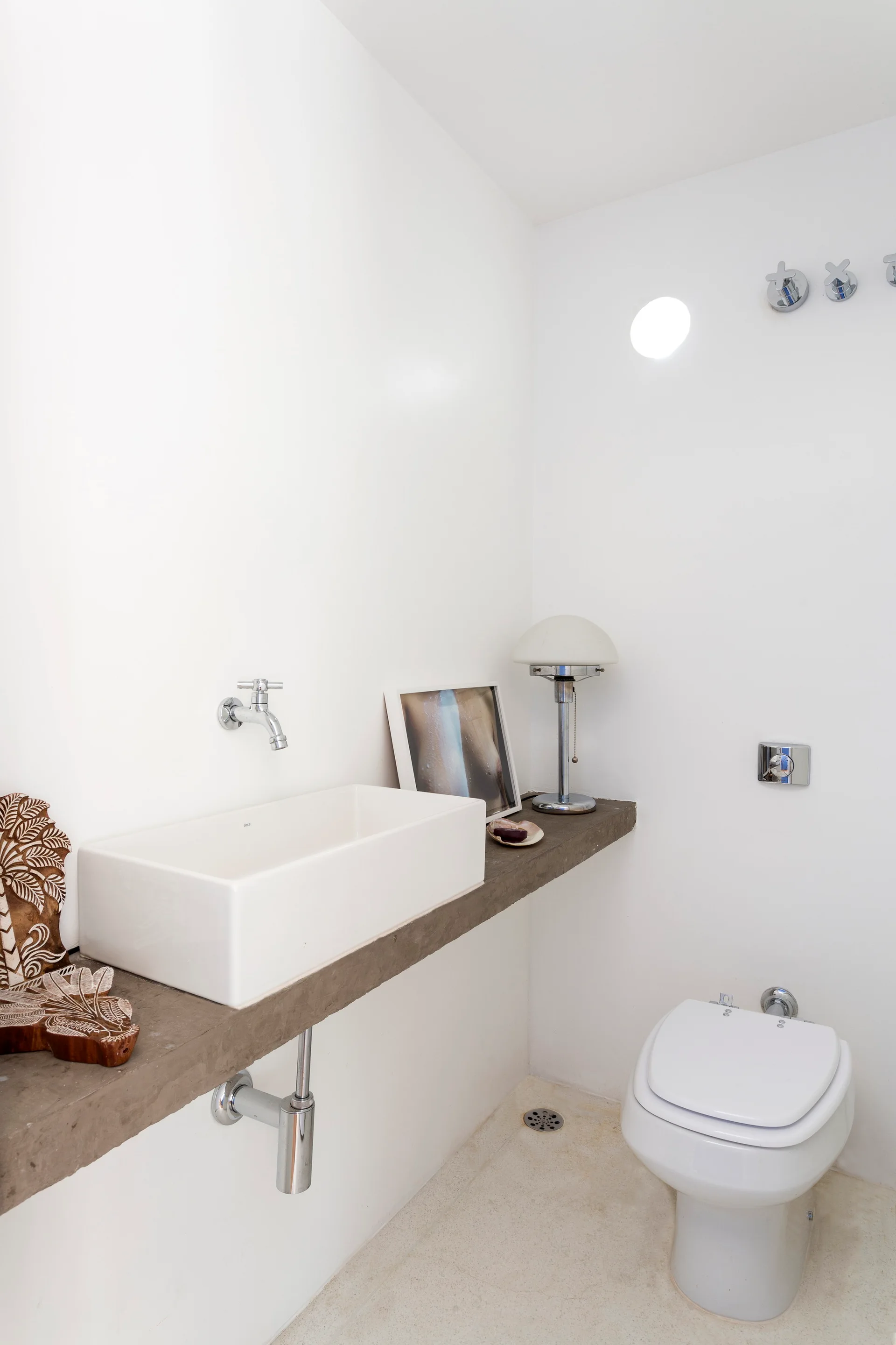 A minimalist white bathroom with a thick concrete floating vanity, rectangular vessel sink, wall-mounted faucet, and a toilet.