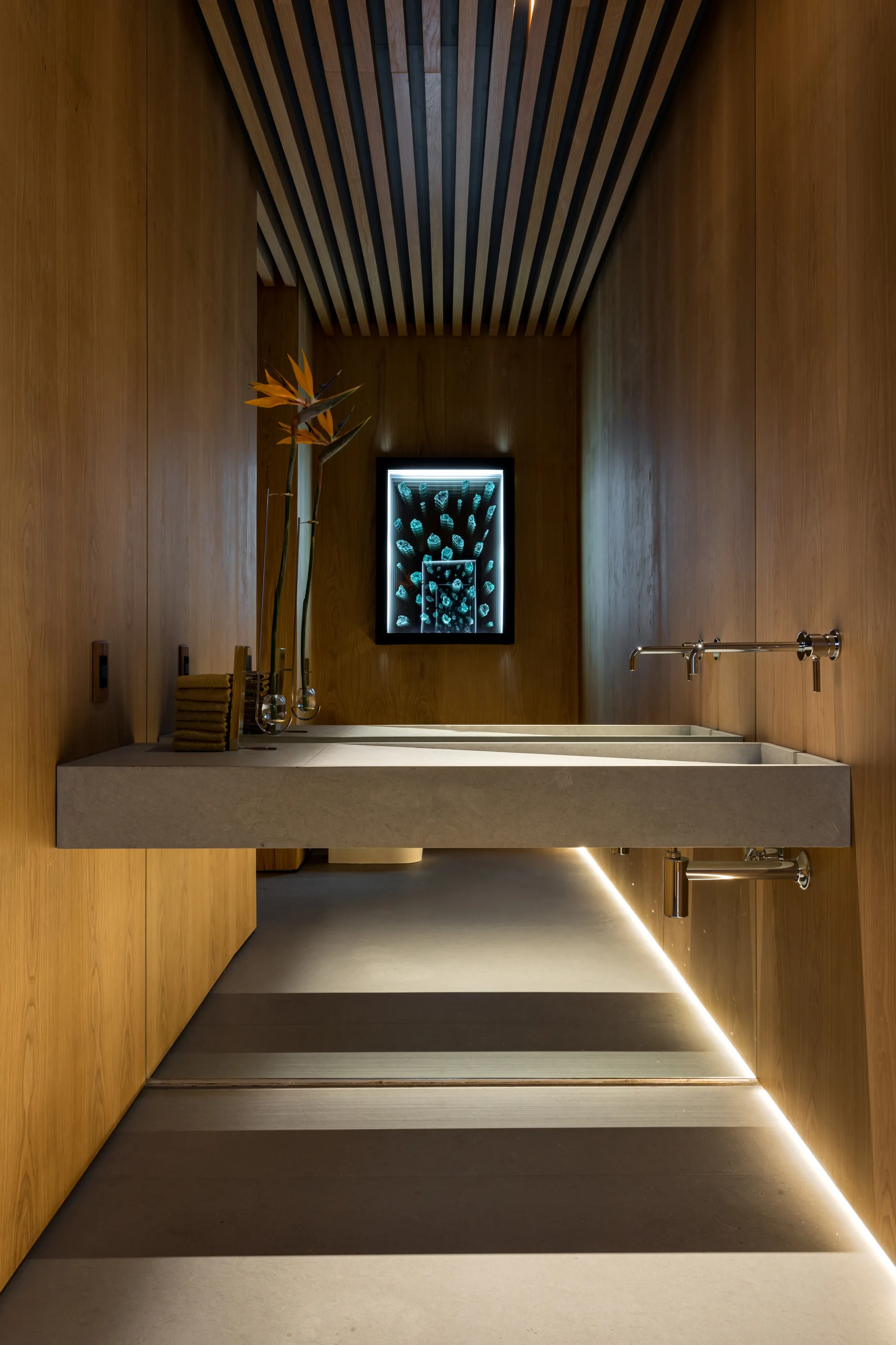 A minimalist bathroom features warm wood paneled walls, a slatted ceiling, and a long floating concrete double vanity with chrome wall-mounted faucets.