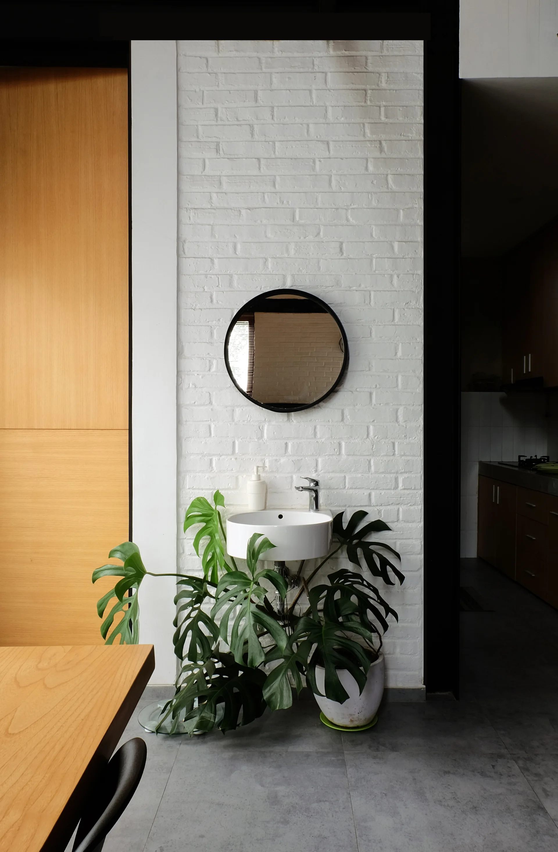 A minimalist white sink and round black mirror are mounted on a white brick wall, with a large potted monstera plant softening the space below.