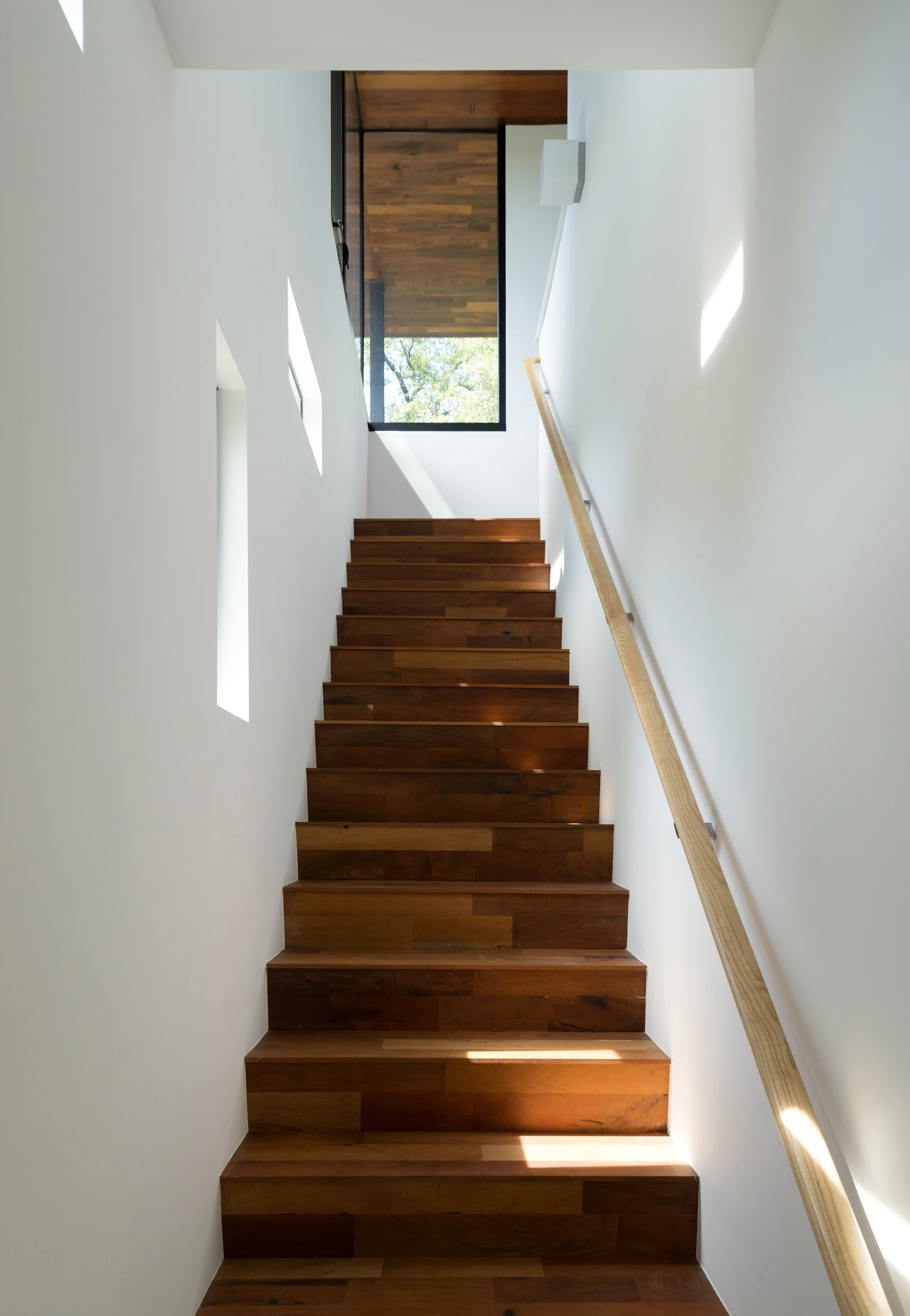 A modern wooden staircase with rich brown treads ascends between smooth white walls, featuring vertical window slits and a light wood handrail.