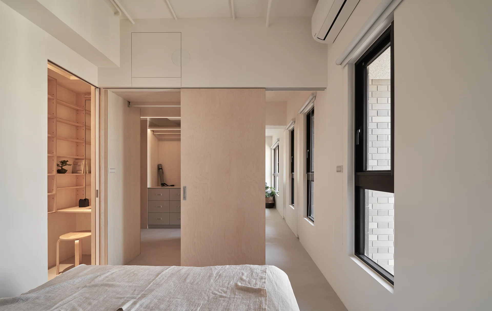 A minimalist white bedroom featuring a large light wood sliding door, built-in shelving, and tall windows along the wall.