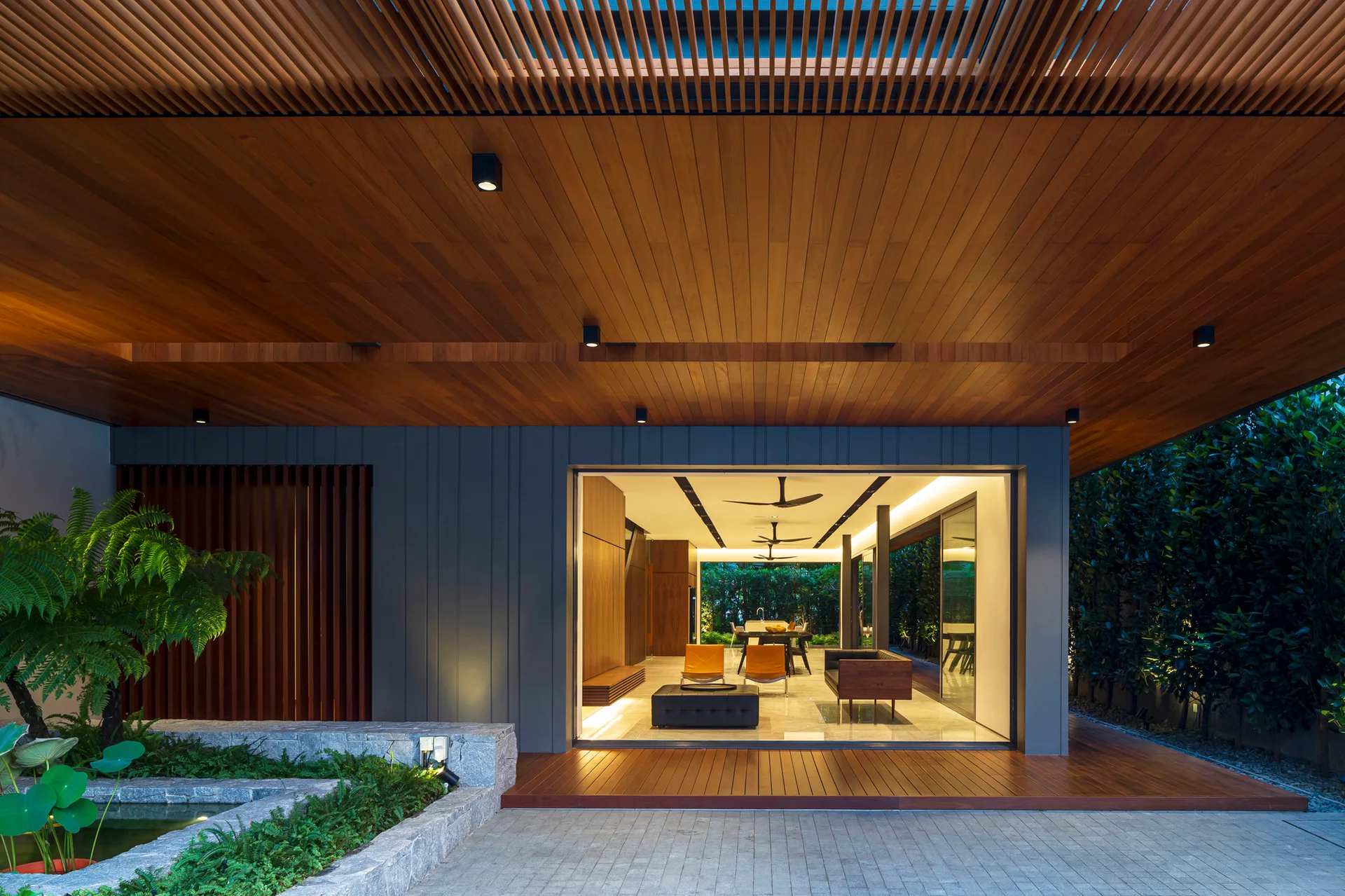 A covered modern patio with a slatted wood ceiling features a stone water pond and opens up to an interior living room with orange chairs.