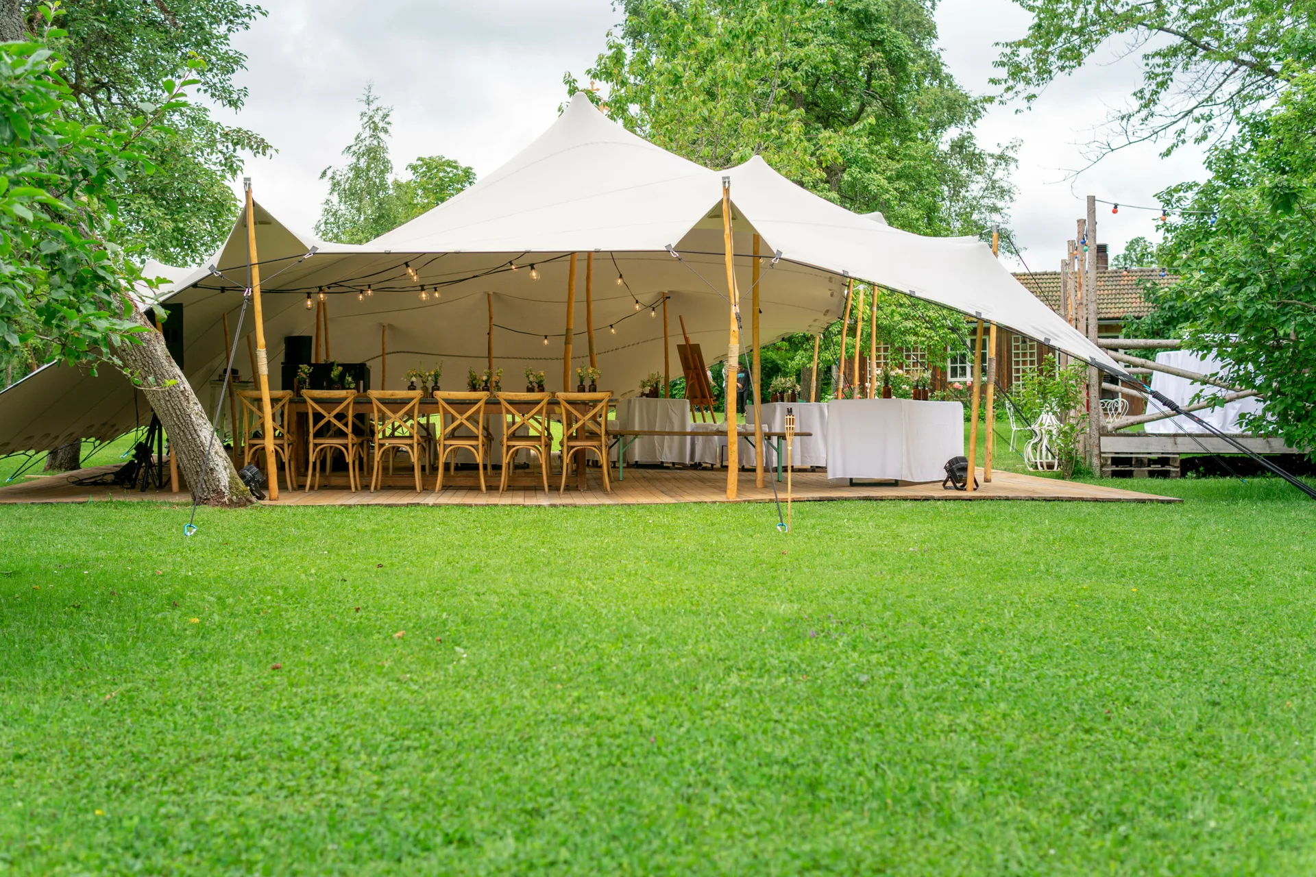 A large white stretch tent stands over a wooden deck with rustic wooden chairs and dining tables arranged on a green lawn.