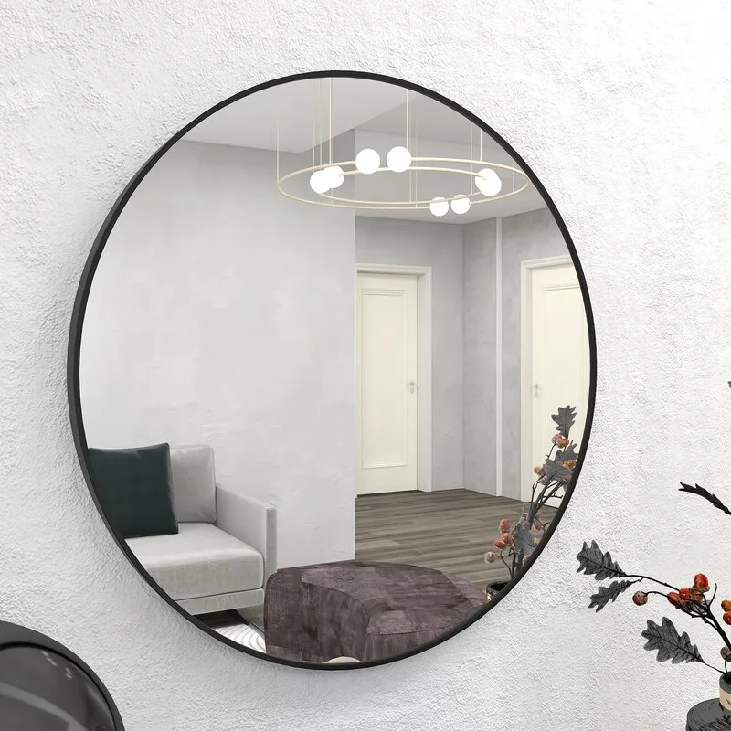 A large circular mirror with a thin black frame hangs on a textured white wall, reflecting a minimalist living room and modern ring light.