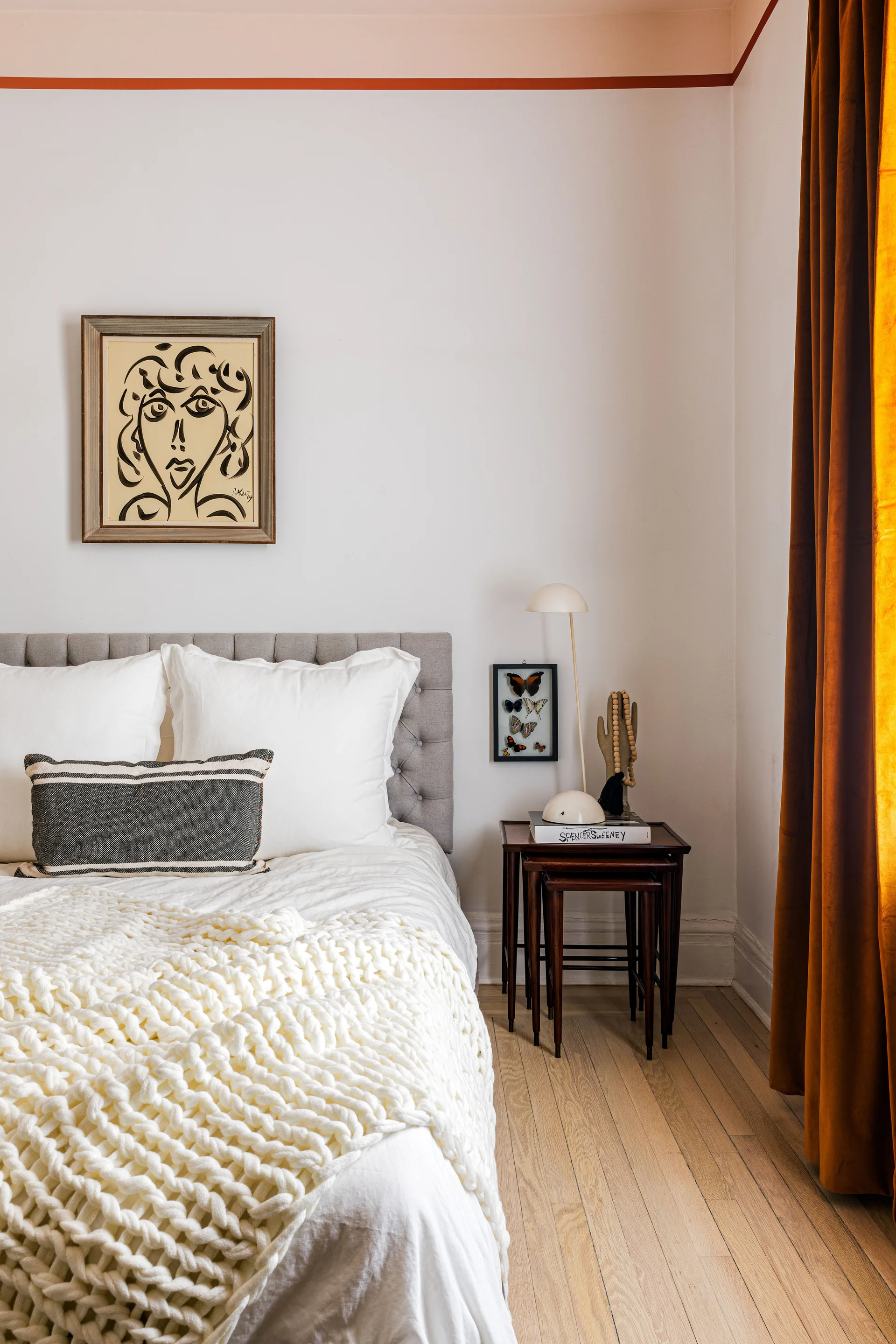 A bedroom corner features a grey tufted headboard, white bedding with a chunky knit throw, wooden nesting tables, and amber velvet curtains.