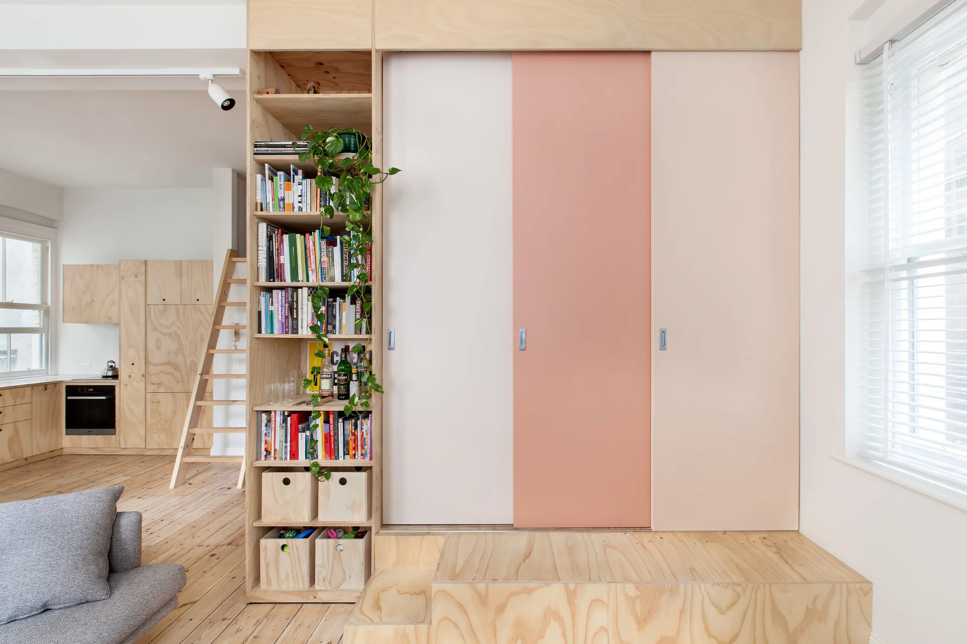 A built-in plywood wardrobe features pastel pink and cream sliding doors on a raised wooden platform next to a tall, integrated bookshelf.