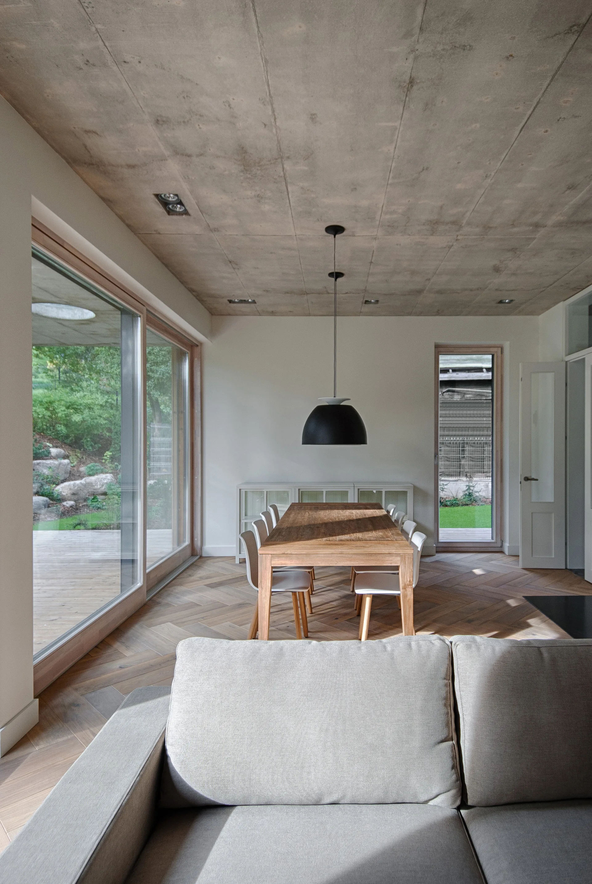 A modern dining room features a wooden table, white chairs, a black dome pendant light, an exposed concrete ceiling, and herringbone wood floors.