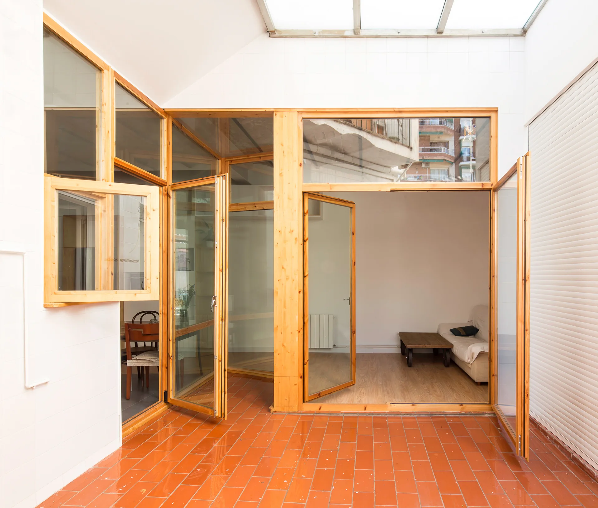 A bright sunroom featuring orange terracotta floor tiles, a translucent skylight, and pine wood-framed glass doors leading into a living room.