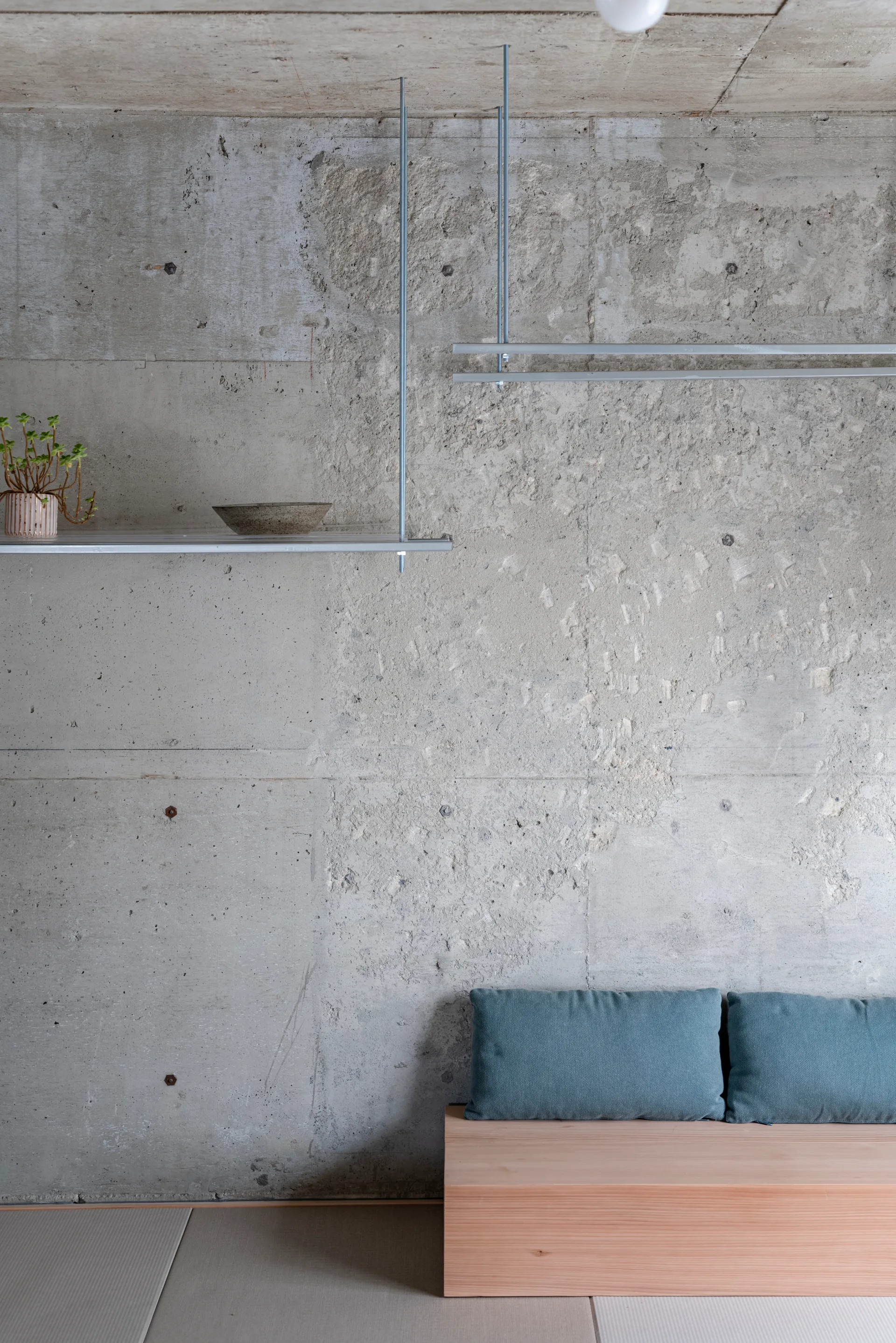 A light wood bench with blue cushions sits against an exposed concrete wall under a suspended metal shelf.