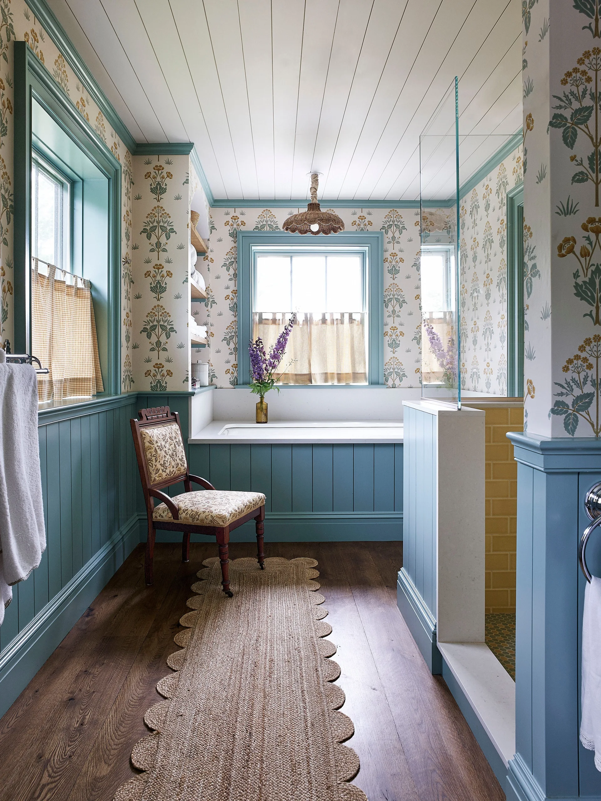 A cottage bathroom features teal wainscoting, floral wallpaper, a built-in bathtub under a window, and a scalloped woven runner on wood floors.