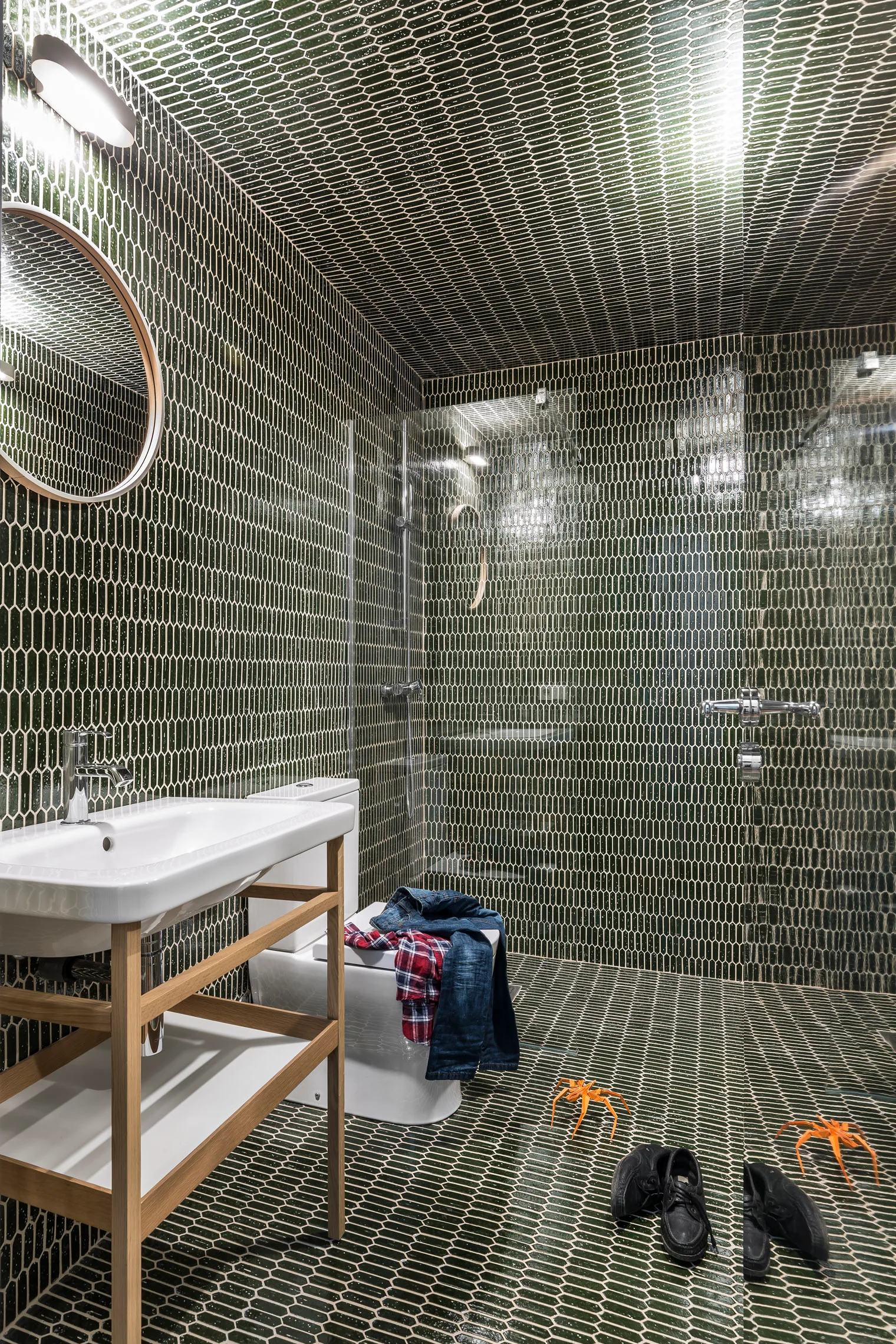A bathroom covered in dark green hexagonal tiles across the walls and ceiling features an open light wood vanity with a white ceramic sink.
