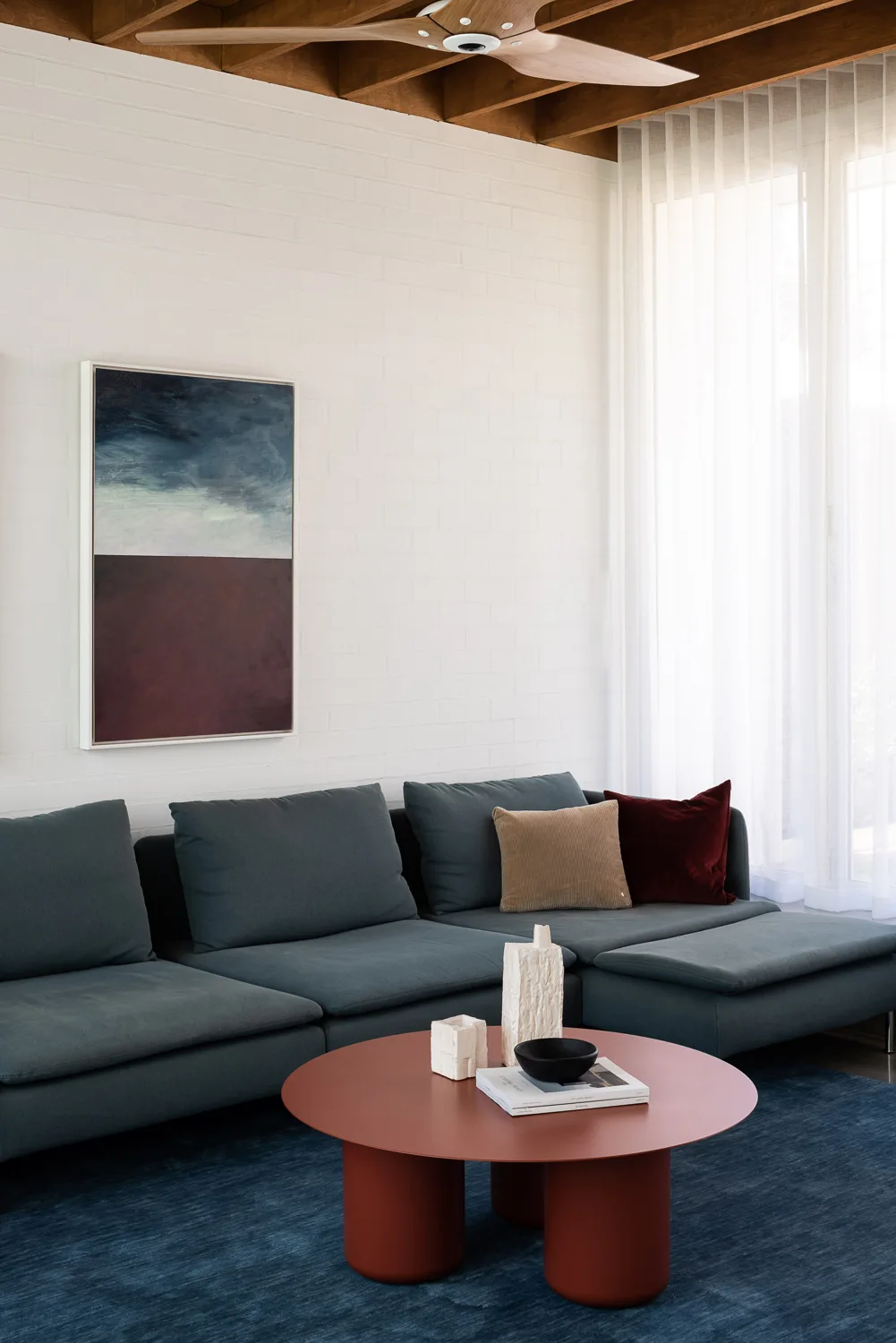 A modern living room features a teal sectional sofa, a round terracotta coffee table, a white brick wall, and exposed wood ceiling beams.