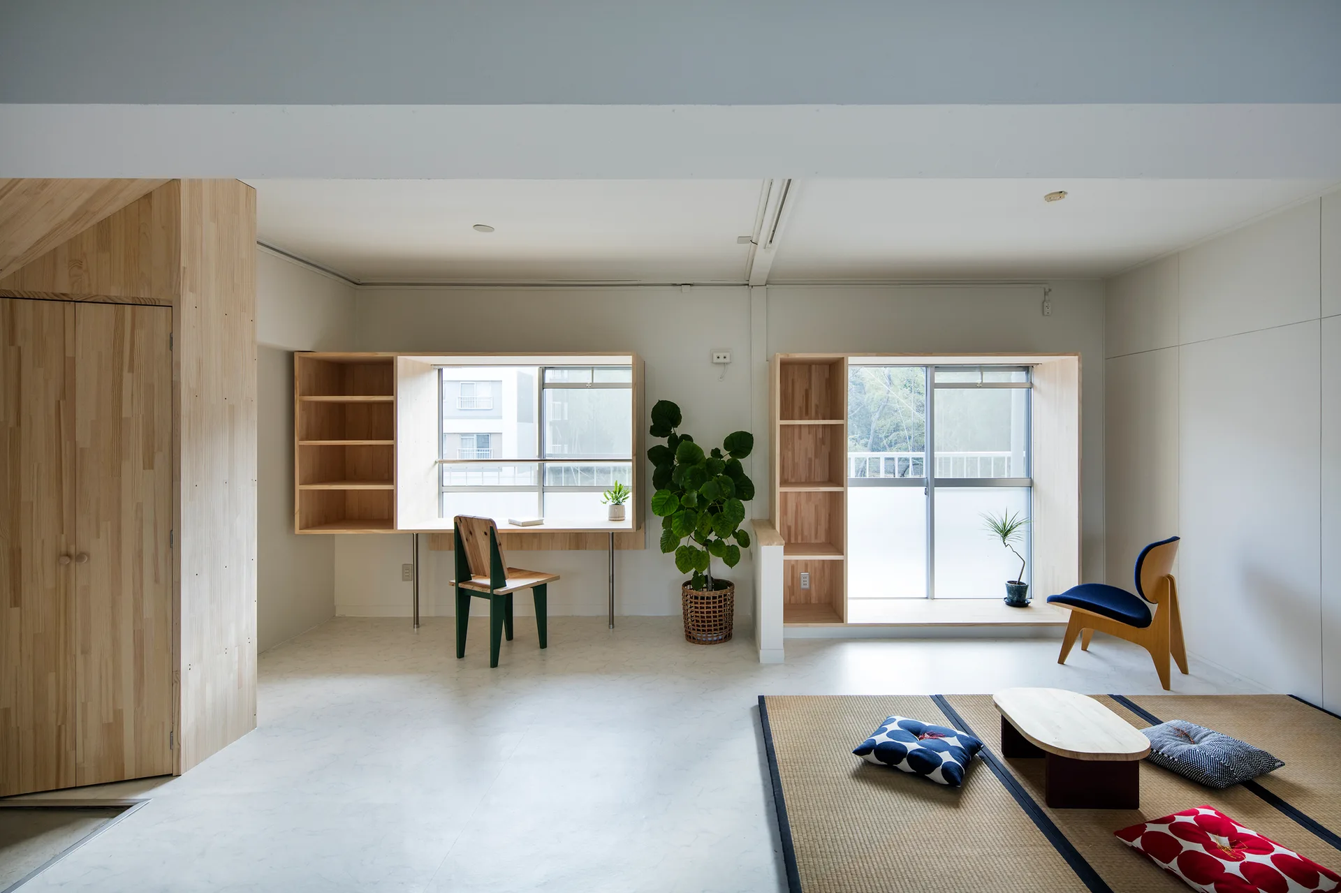 A bright room featuring built-in wooden desks around large windows, a green chair, and a traditional tatami floor mat with cushions.