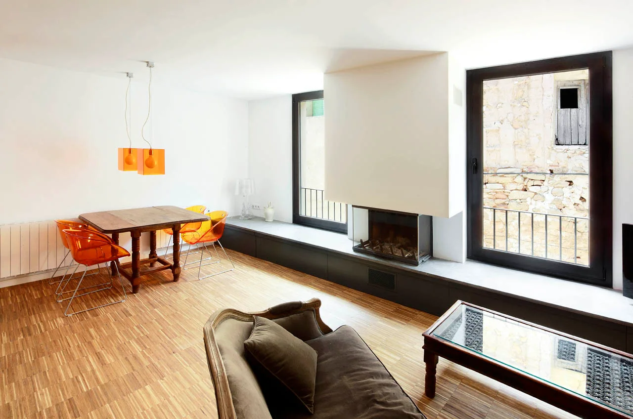 An open-plan living and dining room with white walls, parquet floors, orange dining chairs, and a fireplace set into a long low bench.