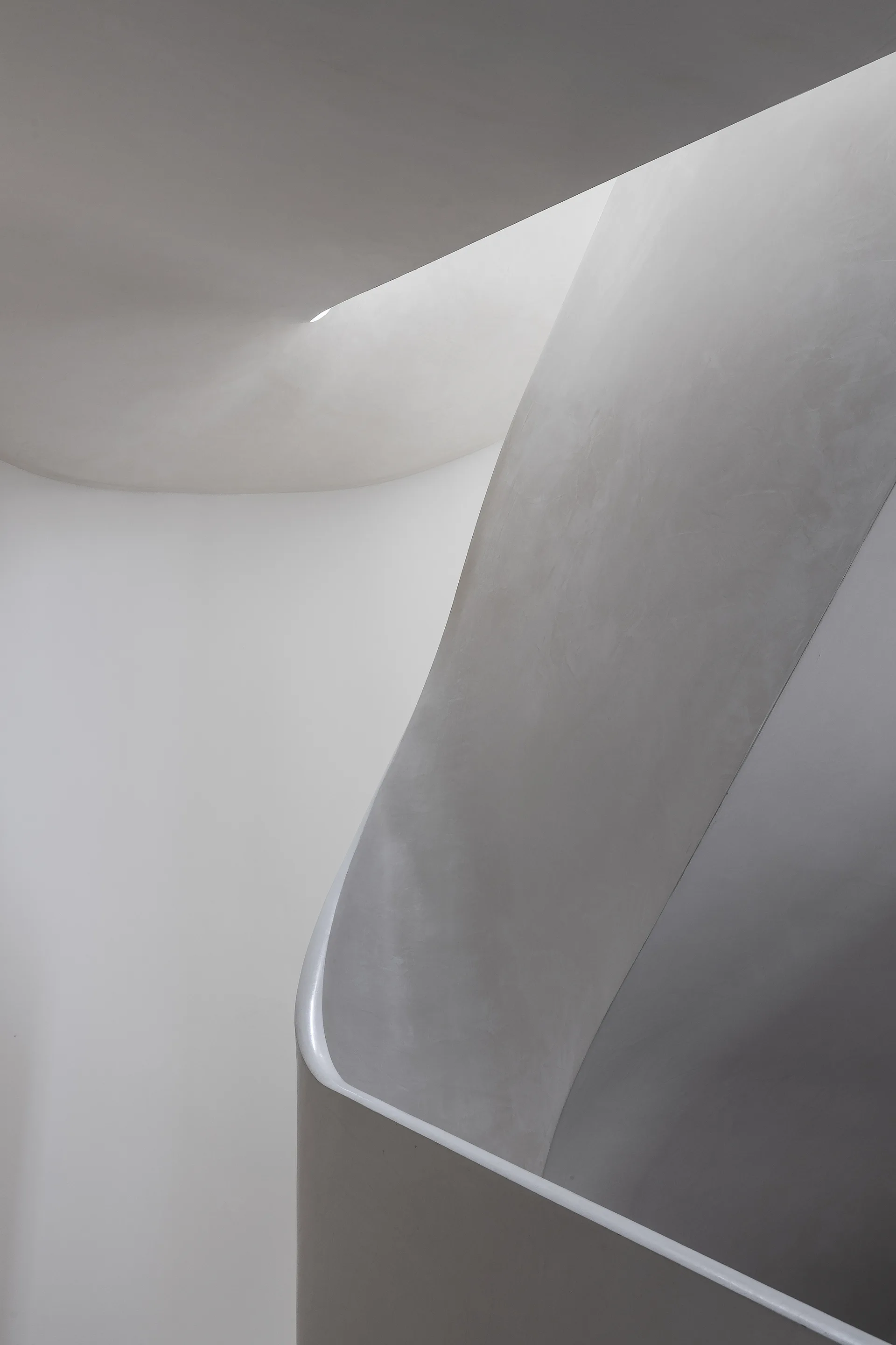 A minimalist curved staircase features smooth off-white plaster walls, a seamless handrail, and a recessed linear light along the upper ceiling curve.