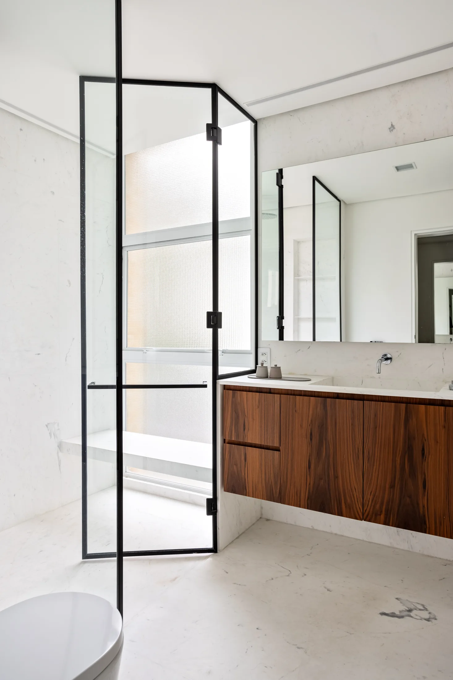 A minimalist bathroom features a floating dark wood vanity, white marble tile walls, and a black-framed glass shower door next to a large mirror.