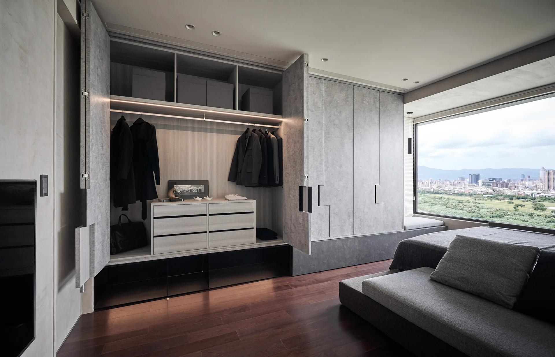 A modern dressing room featuring a built-in gray wardrobe with open shelves, integrated lighting, a chest of drawers, and a large window.