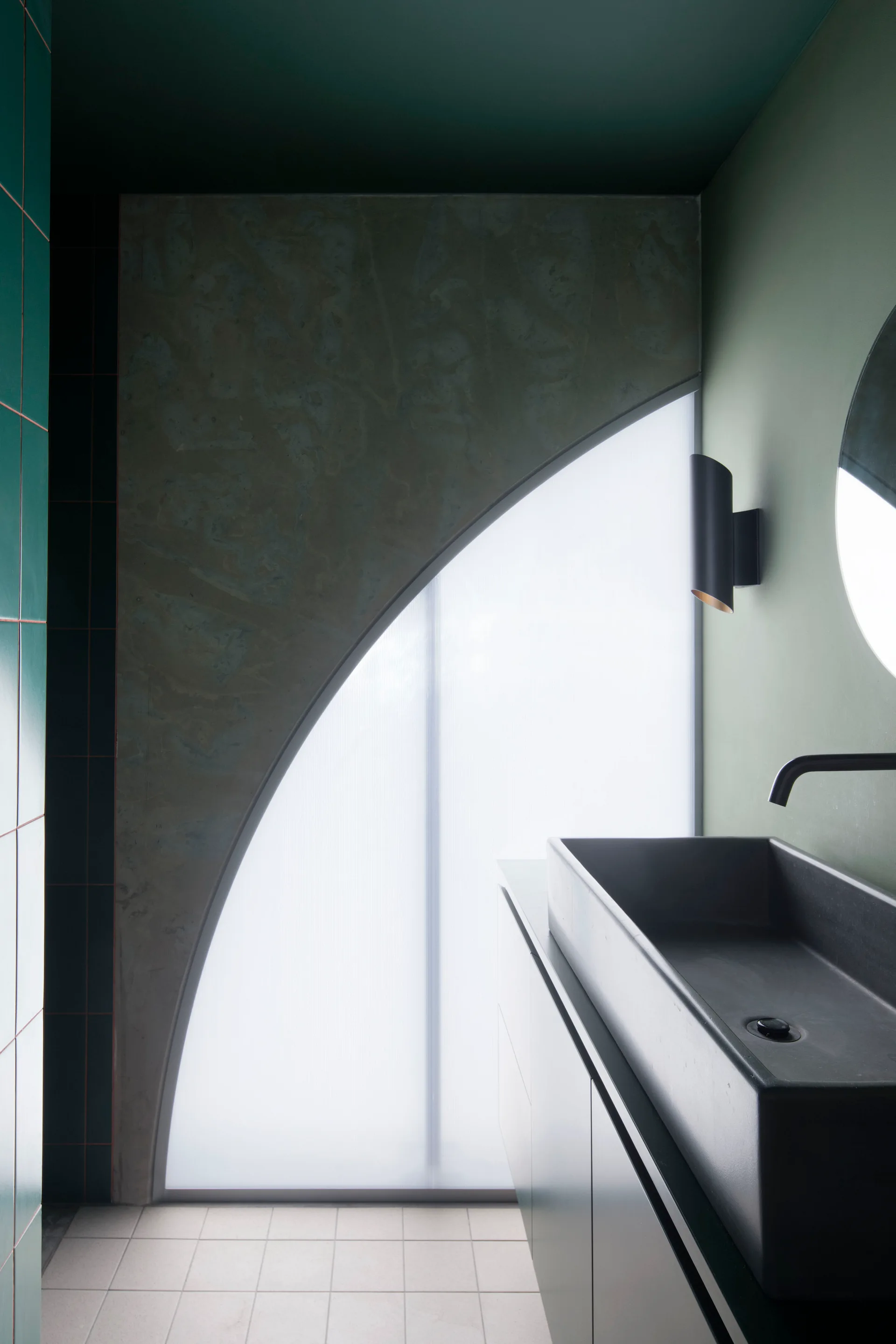 A minimalist bathroom vanity area with a black rectangular vessel sink, dark green walls, and a large arched frosted glass window.