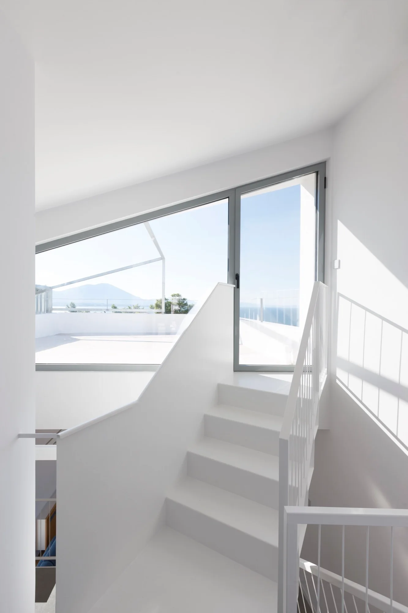 A minimalist white indoor staircase ascends toward a large angular glass door and window that opens onto a sunny outdoor rooftop terrace.