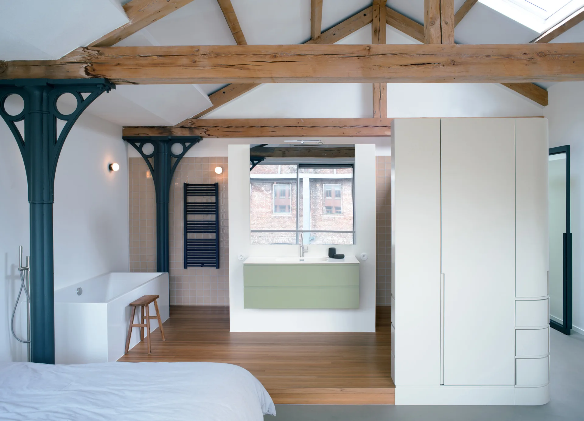 An open-concept loft bedroom features exposed wooden rafters, a built-in white bathtub, a green floating vanity, and a large wardrobe.