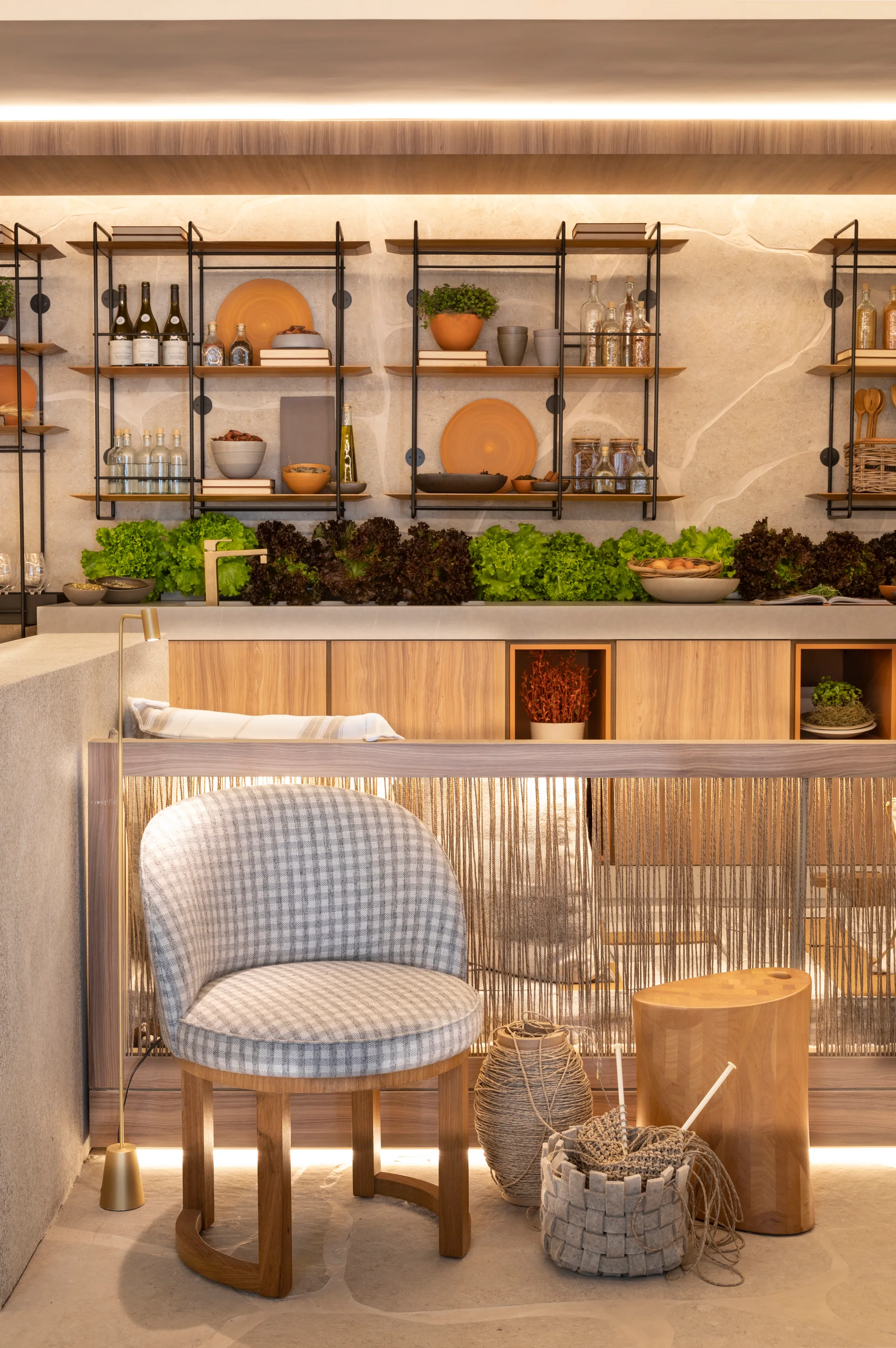 A plaid armchair stands near a wooden stool in front of a warm-toned kitchen with wood cabinets, open black metal shelves, and a stone wall.