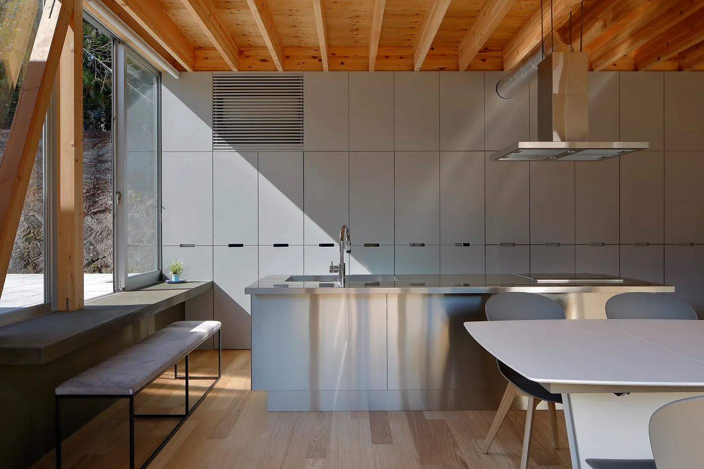 A minimalist kitchen featuring a stainless steel island, a wall of matte grey built-in cabinets, and exposed wood ceiling beams.