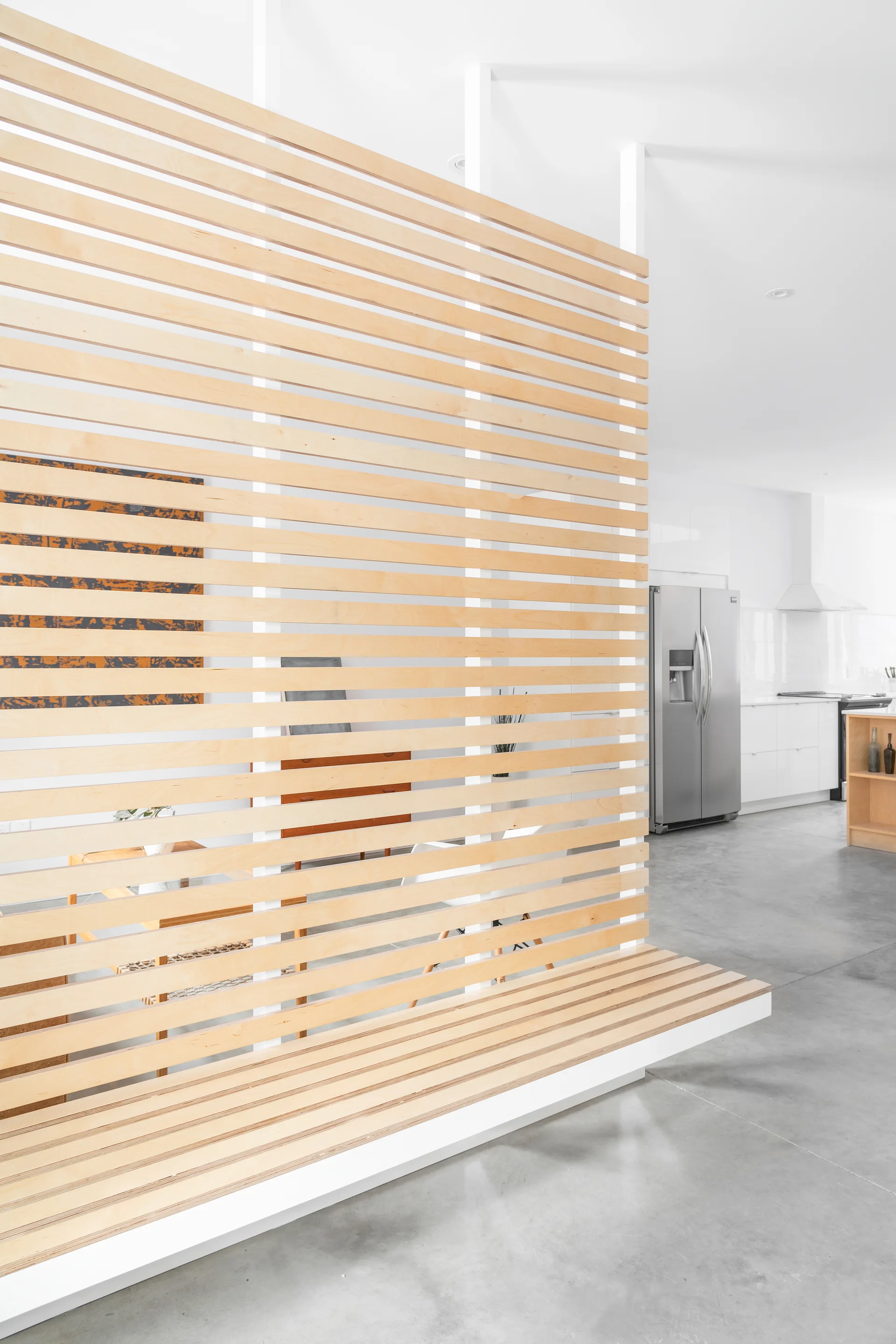 A light wood slatted room divider with an integrated low bench stands on a concrete floor next to an open kitchen with white cabinets.
