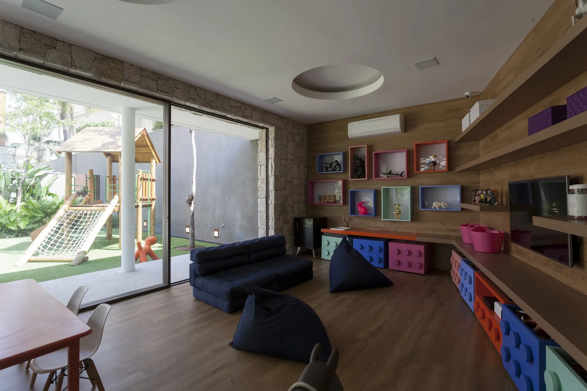 A kids' playroom features wood paneling, colorful toy storage blocks, navy blue bean bags, and sliding glass doors looking out to a playground.