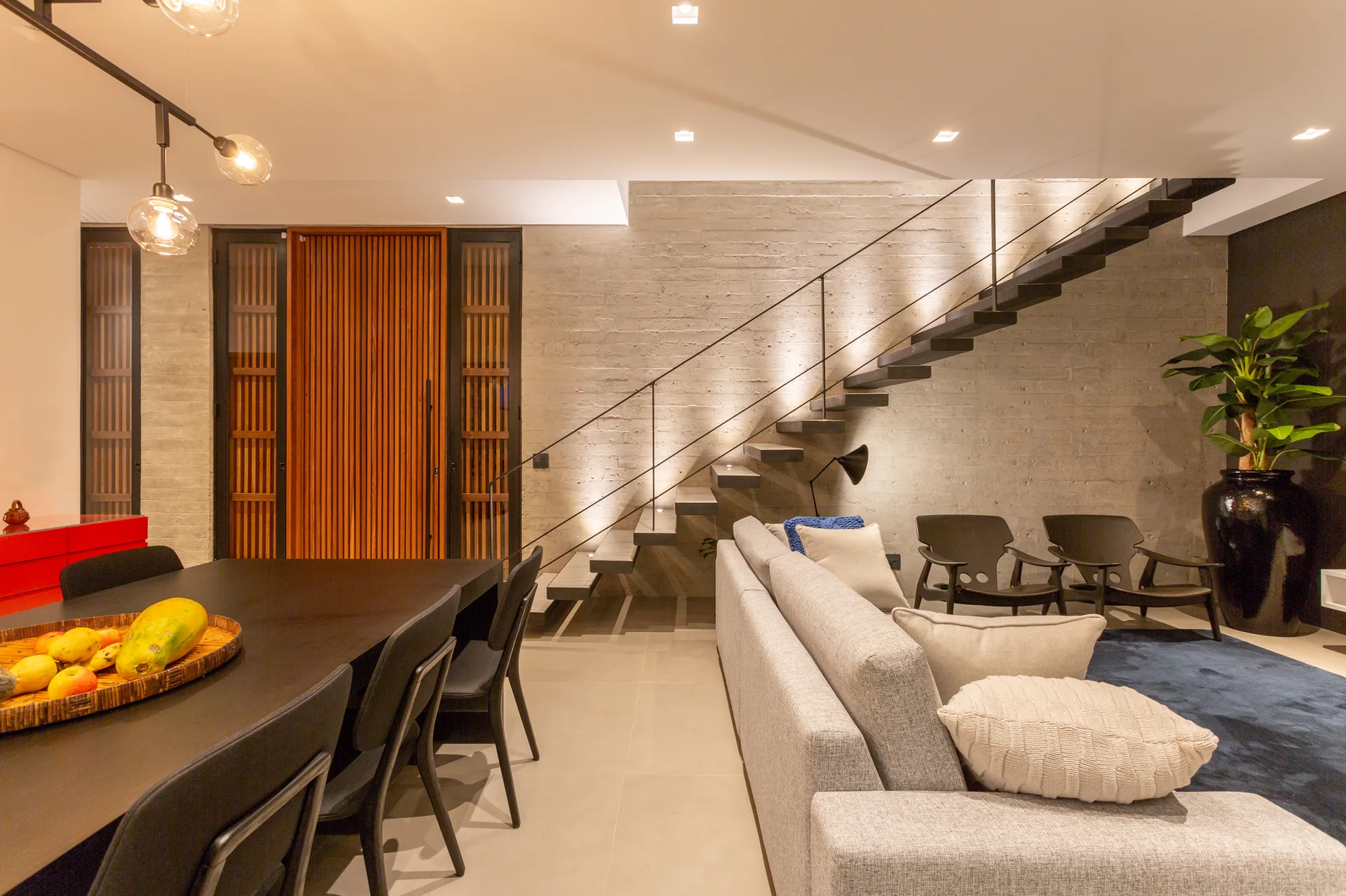 An open-concept living space features a light gray sofa and a black dining table near floating stairs against a textured concrete wall.