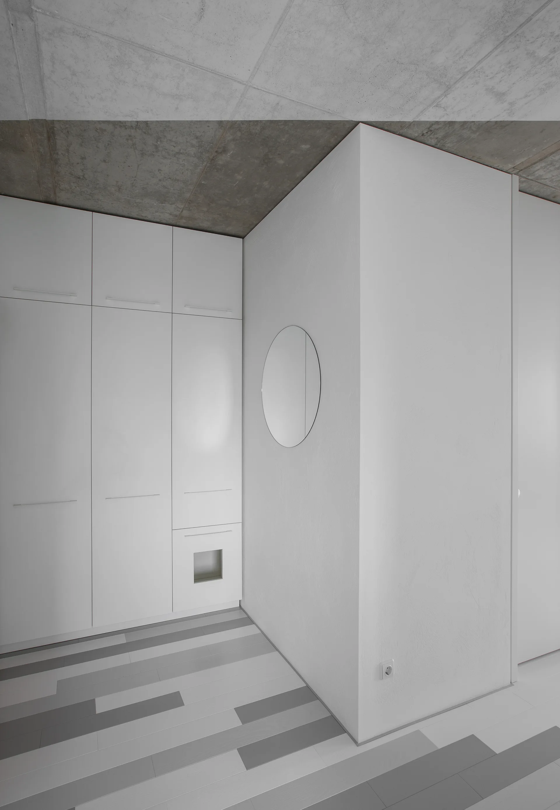 A minimalist hallway with built-in white cabinets, a circular mirror, and multi-tone grey plank flooring beneath an exposed concrete ceiling.