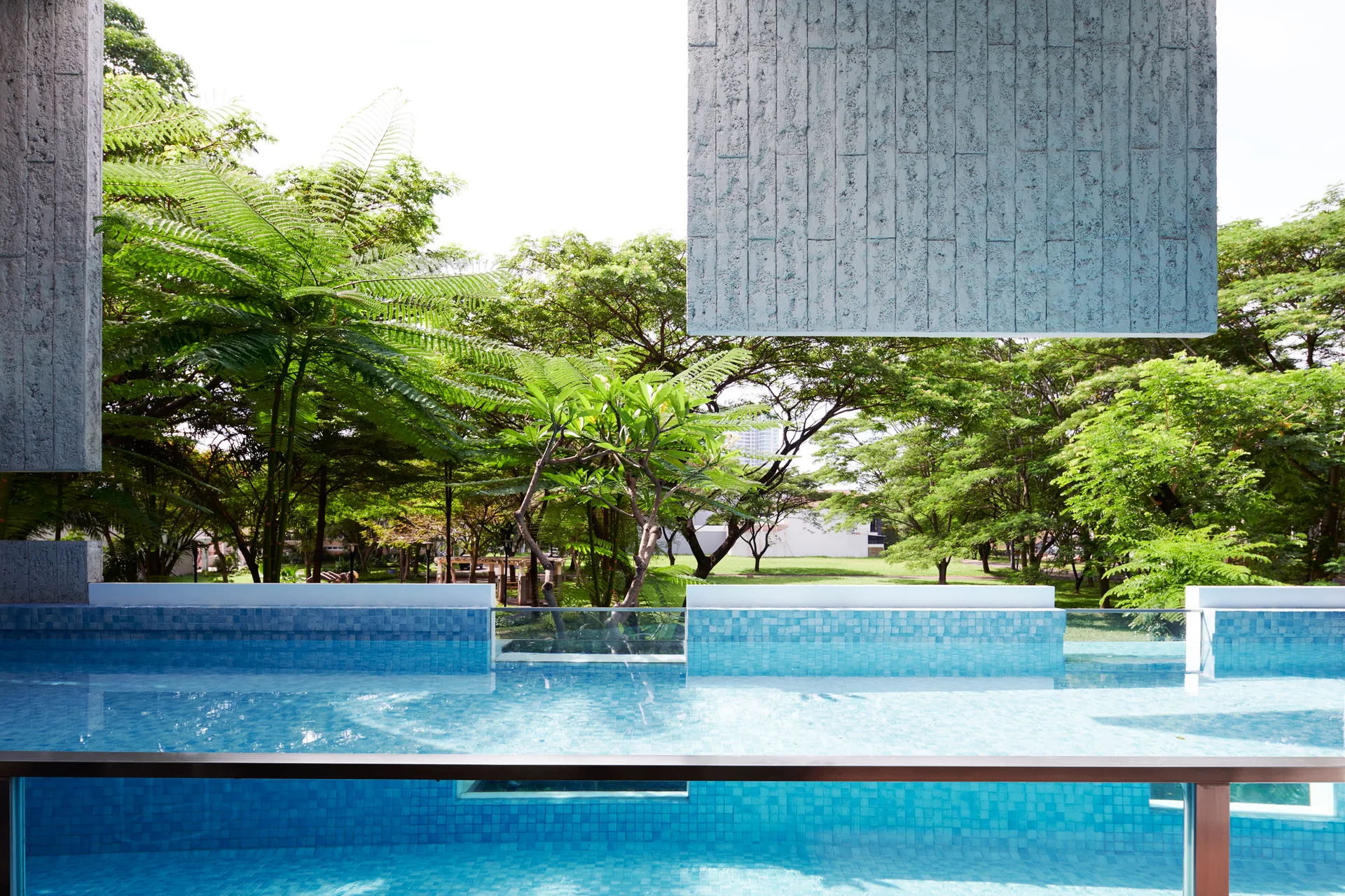 An elevated swimming pool lined with blue mosaic tile and a glass railing overlooks a park with lush green trees.
