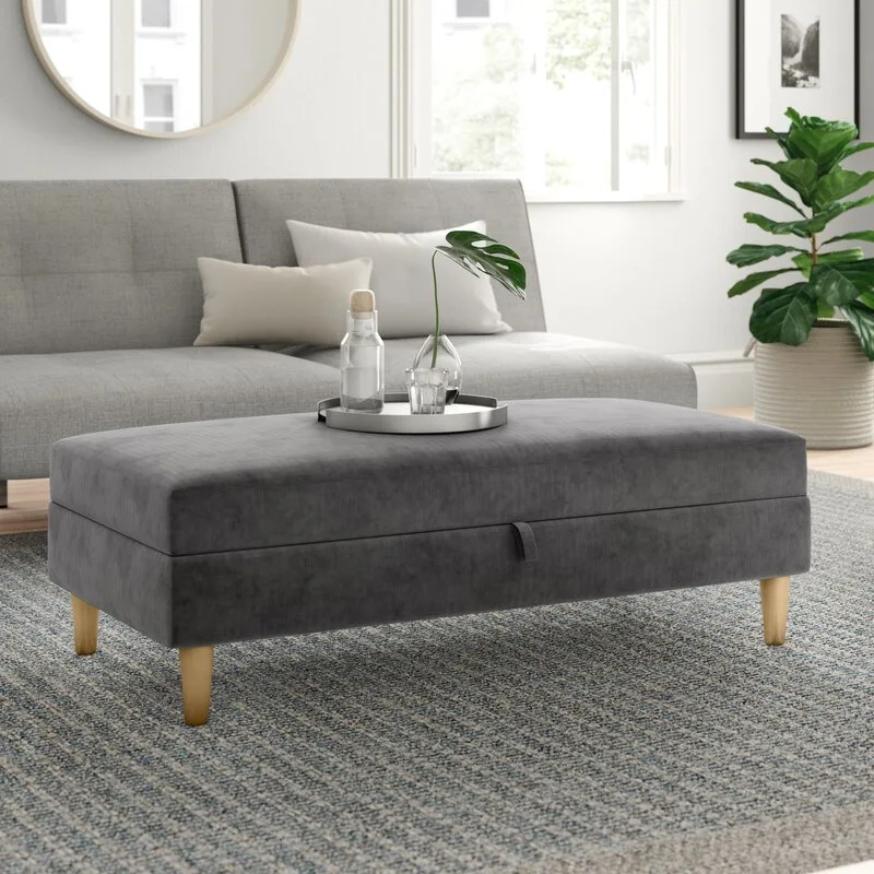 A rectangular gray upholstered storage ottoman with tapered wood legs sits on a textured blue-gray rug in a bright living room.