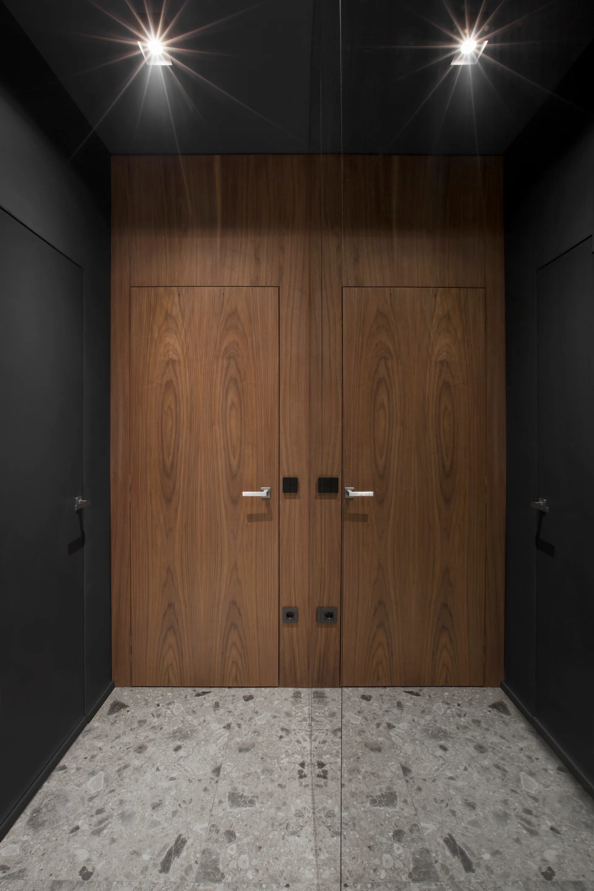 A narrow charcoal gray hallway features a flush wood grain door, matte black walls, and a speckled gray terrazzo floor.
