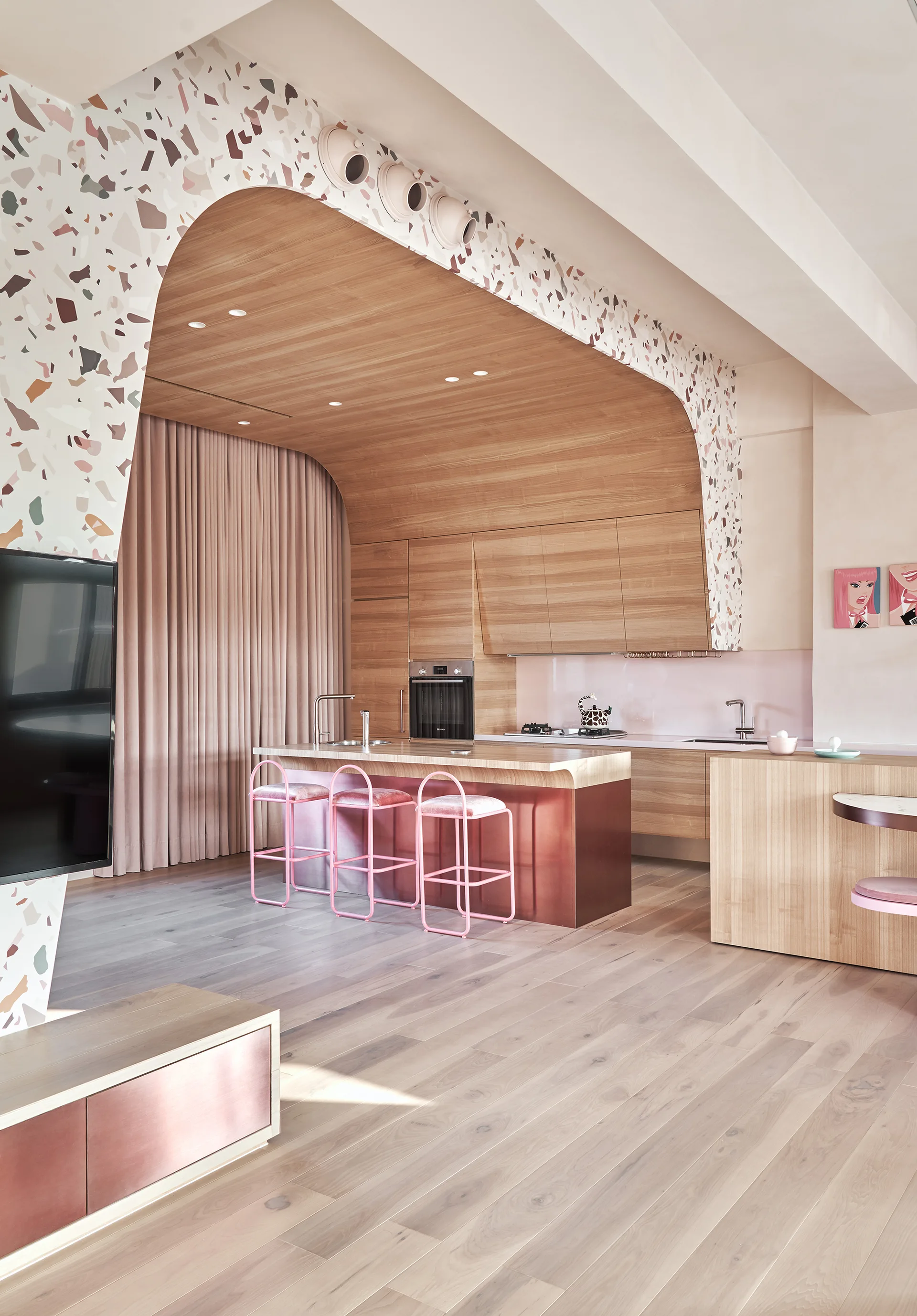 A modern kitchen featuring a wood-clad archway framed in terrazzo, pink metal bar stools at a copper-toned island, and light wood floors.