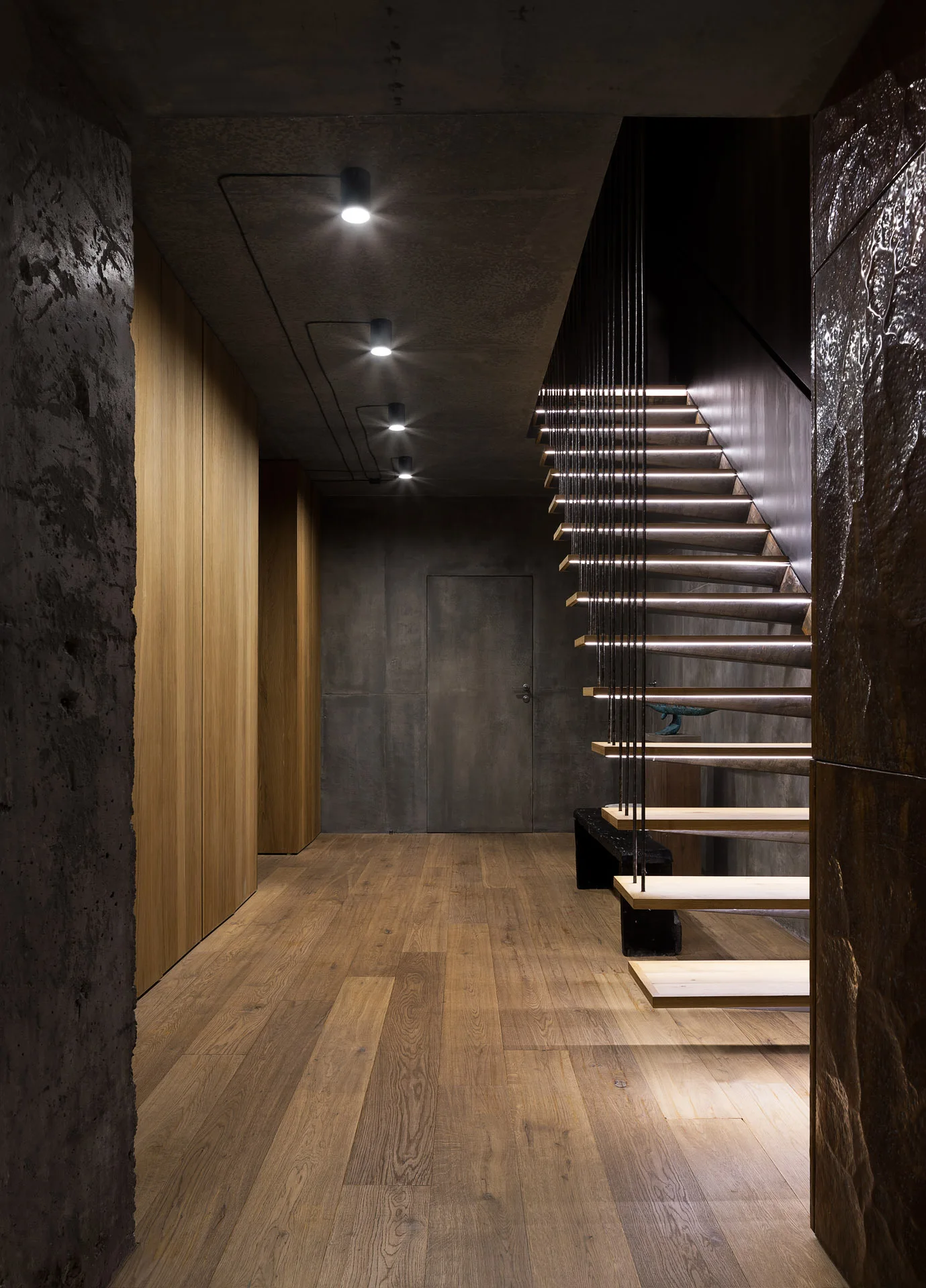 A modern hallway features a floating wood staircase with integrated tread lighting, dark concrete walls, and light oak floorboards.