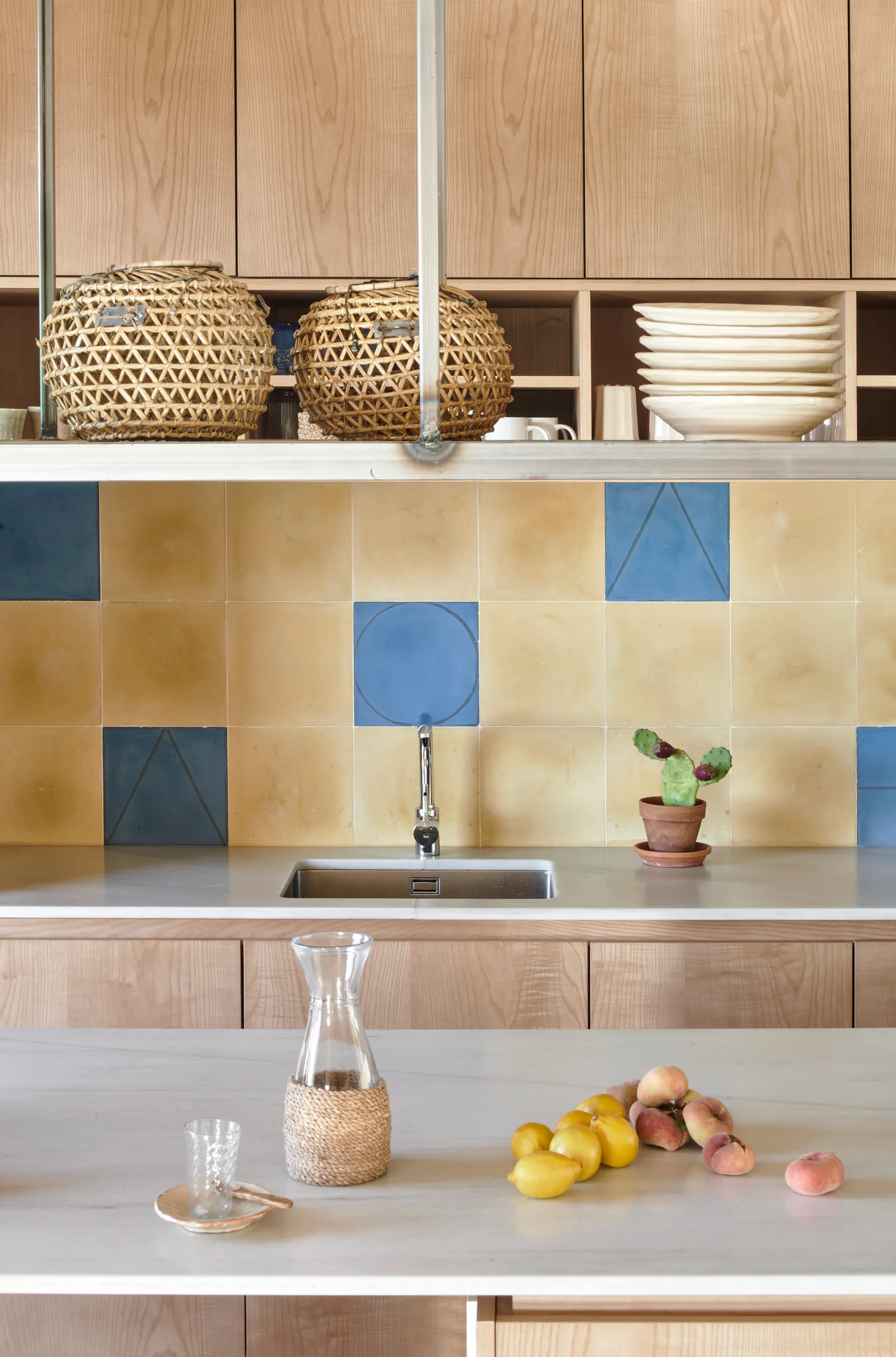 A residential kitchen features flat-front wood cabinets, a yellow and blue geometric tile backsplash, and a marble island with lemons.