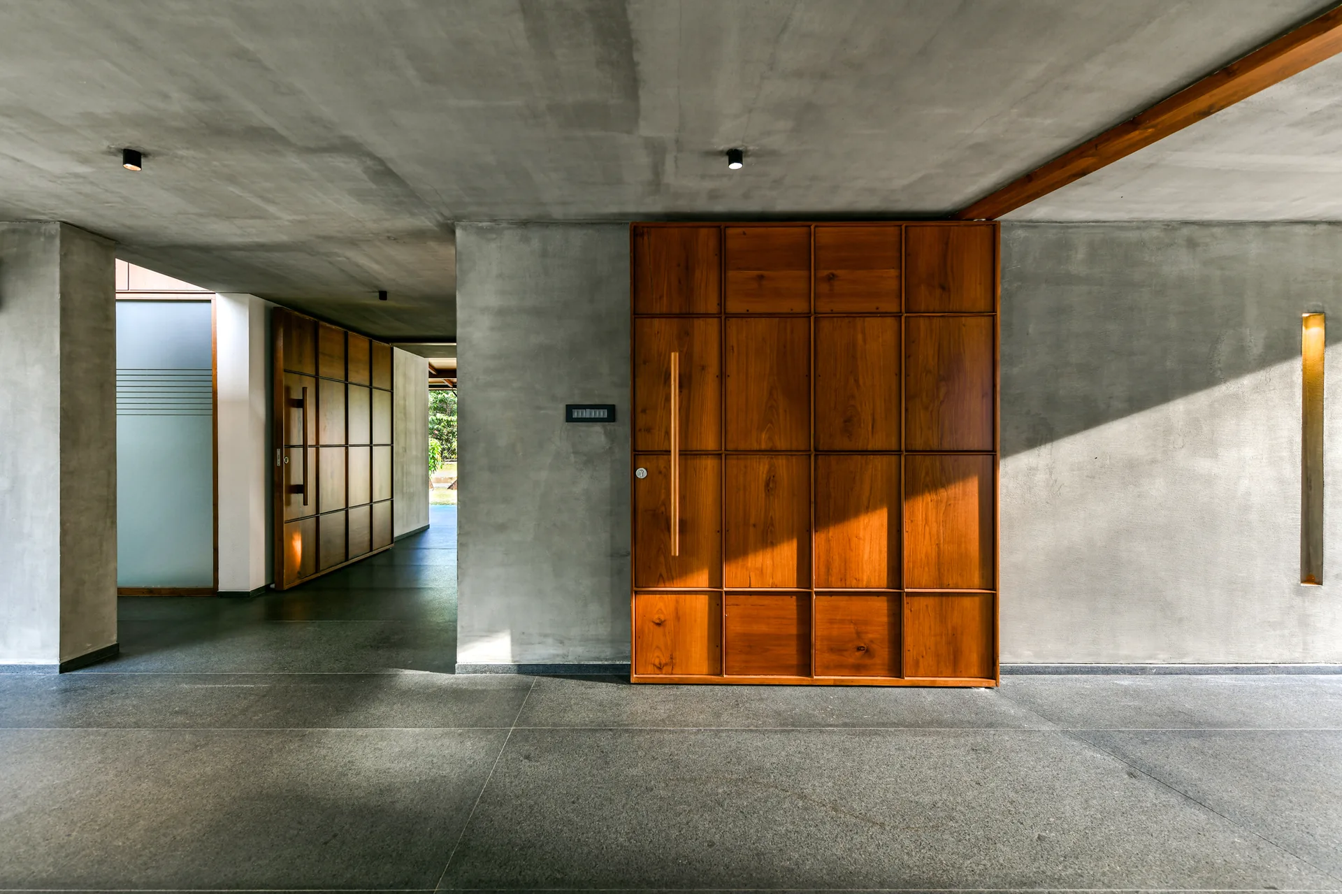 A minimalist entryway features a large grid-patterned wooden door set against smooth, exposed concrete walls and a dark gray stone tile floor.