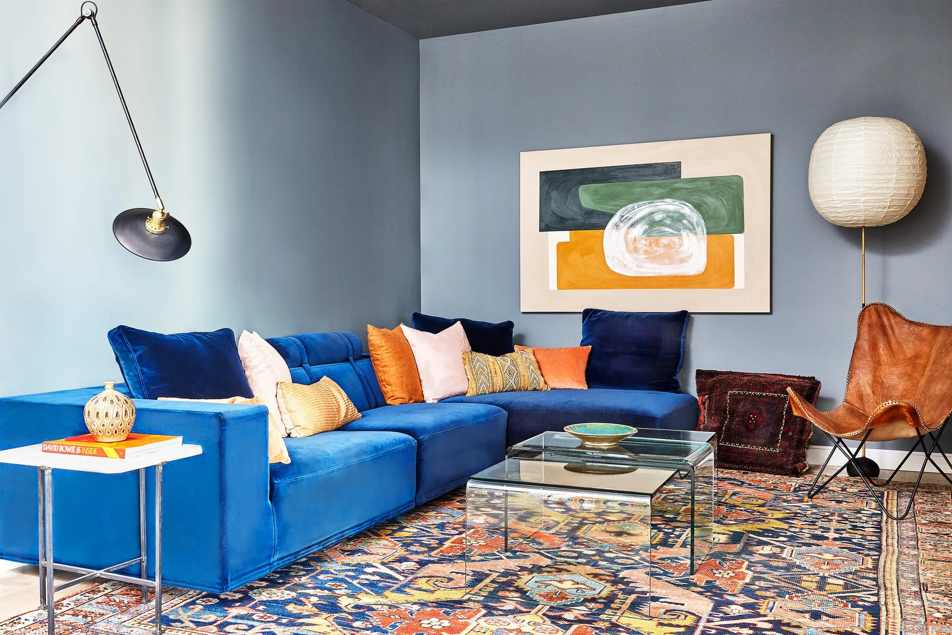 A blue velvet sectional sofa is paired with glass nested coffee tables, a patterned area rug, and an abstract painting against slate gray walls.