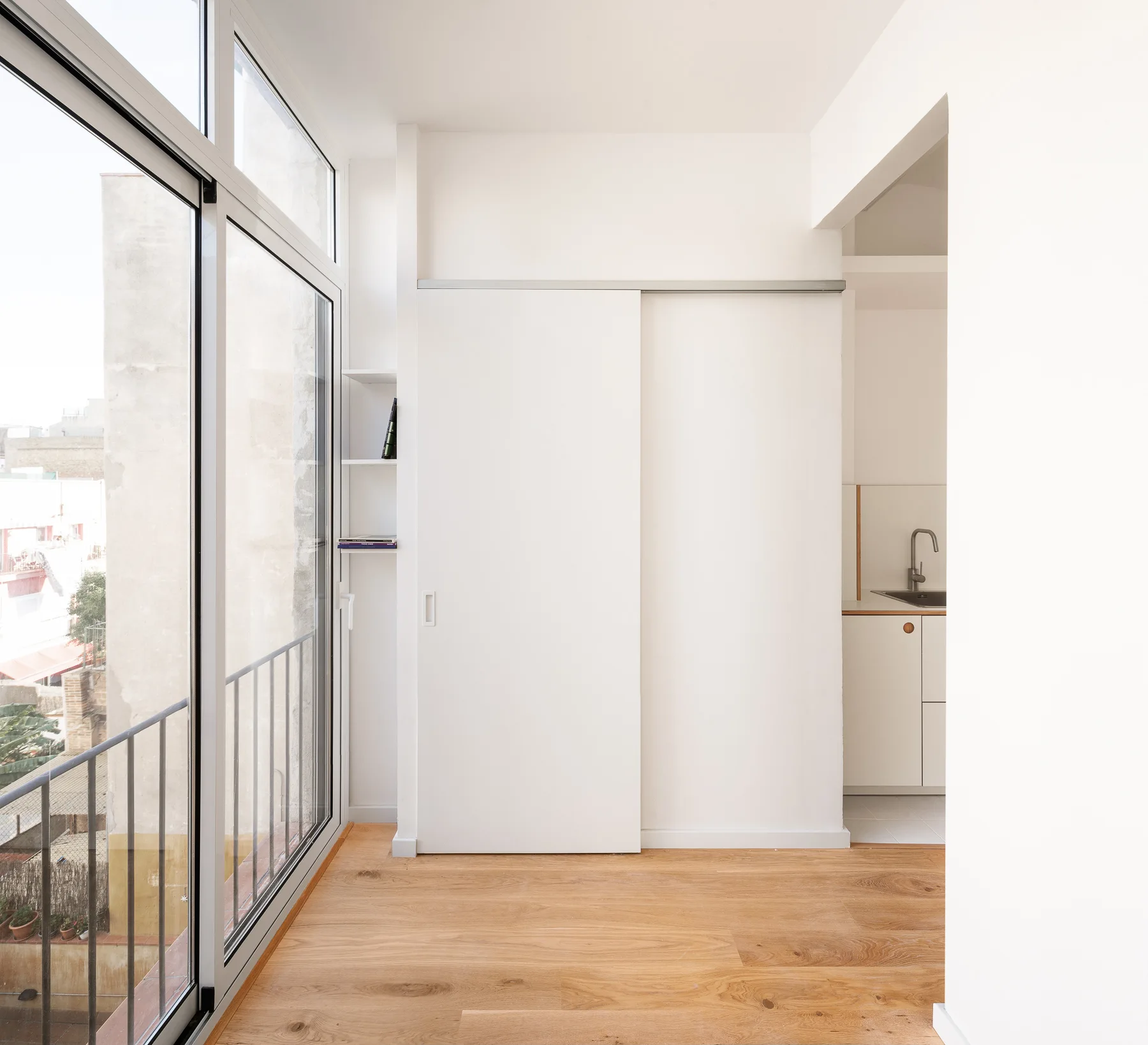 A bright room features a large white sliding partition door next to tall glass windows leading to a balcony, with light wood flooring.