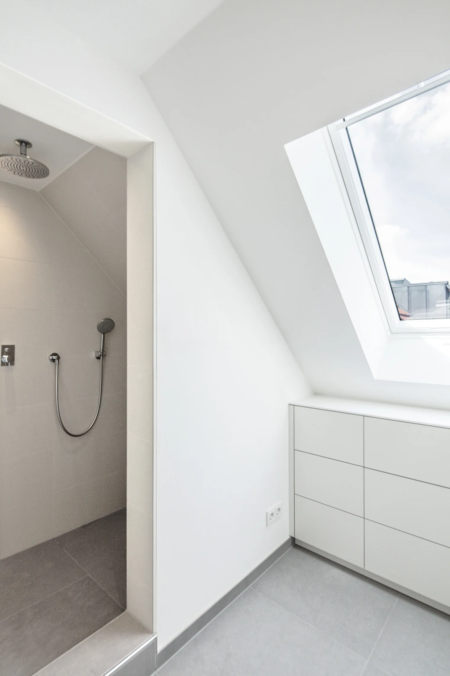 A minimalist attic bathroom featuring grey tiled flooring, a sloped ceiling with a skylight, white built-in cabinetry, and a walk-in shower.