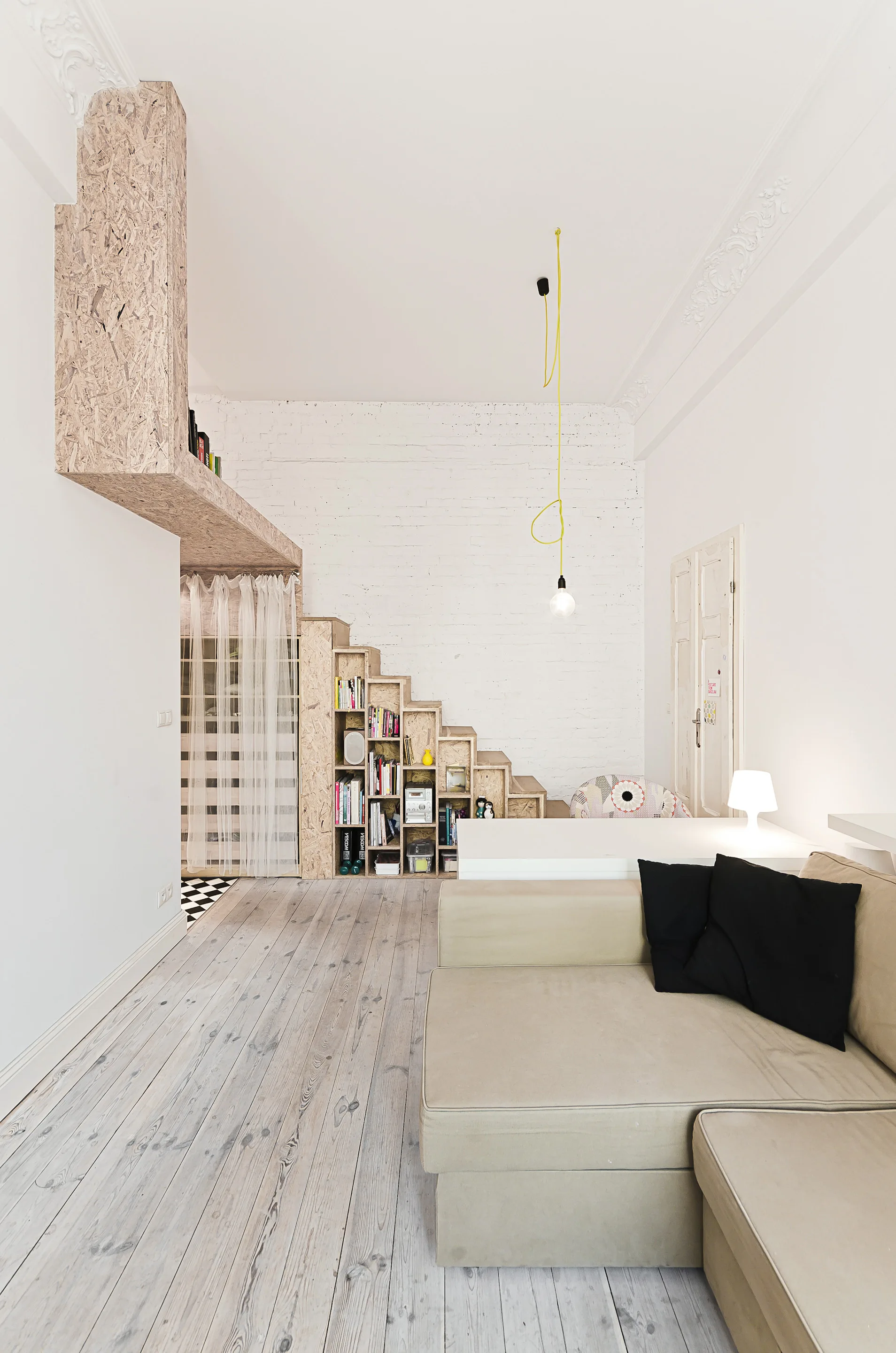 A living area features a tan sectional sofa, light wood floors, a white brick wall, and a staircase-shaped bookcase made of oriented strand board.
