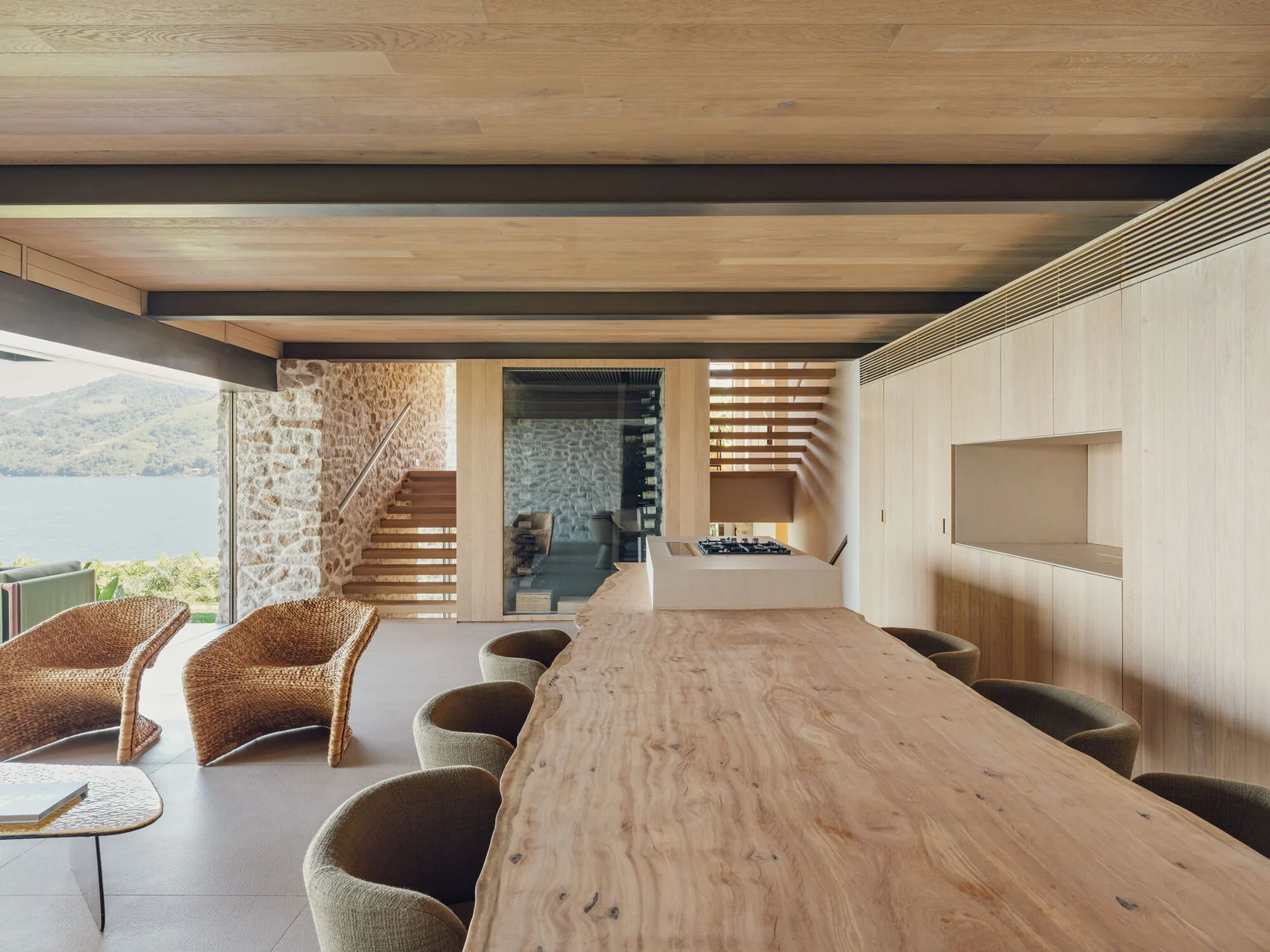 An open-concept dining room features a long live-edge wooden table, a light wood-paneled ceiling with dark beams, and views of a nearby lake.