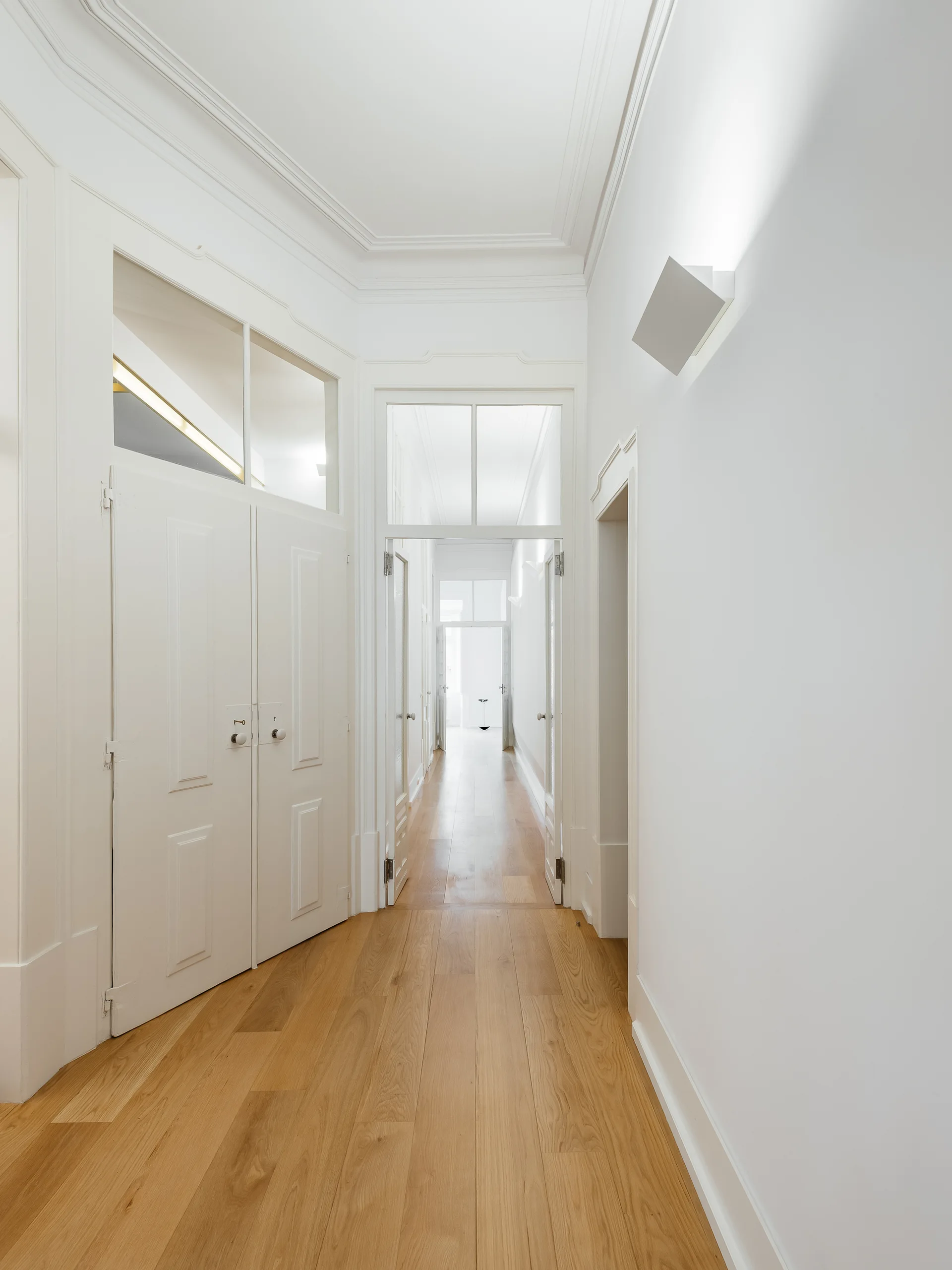 A long white hallway with light oak floors features multiple tall paneled doors with clear glass transom windows above them.