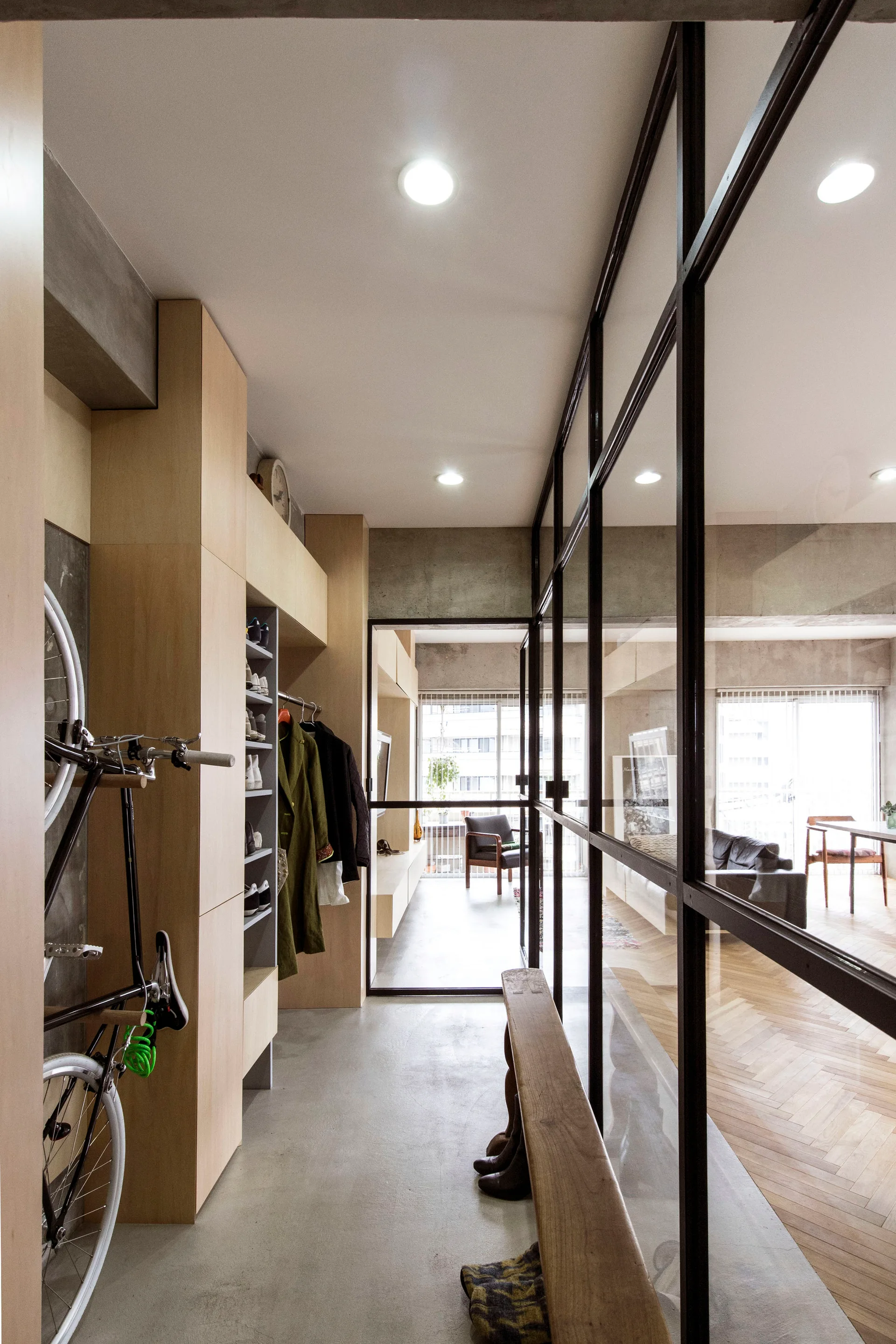A modern entryway features light wood storage cabinets, a wall-mounted bicycle, and a black-framed glass partition looking into a living room.