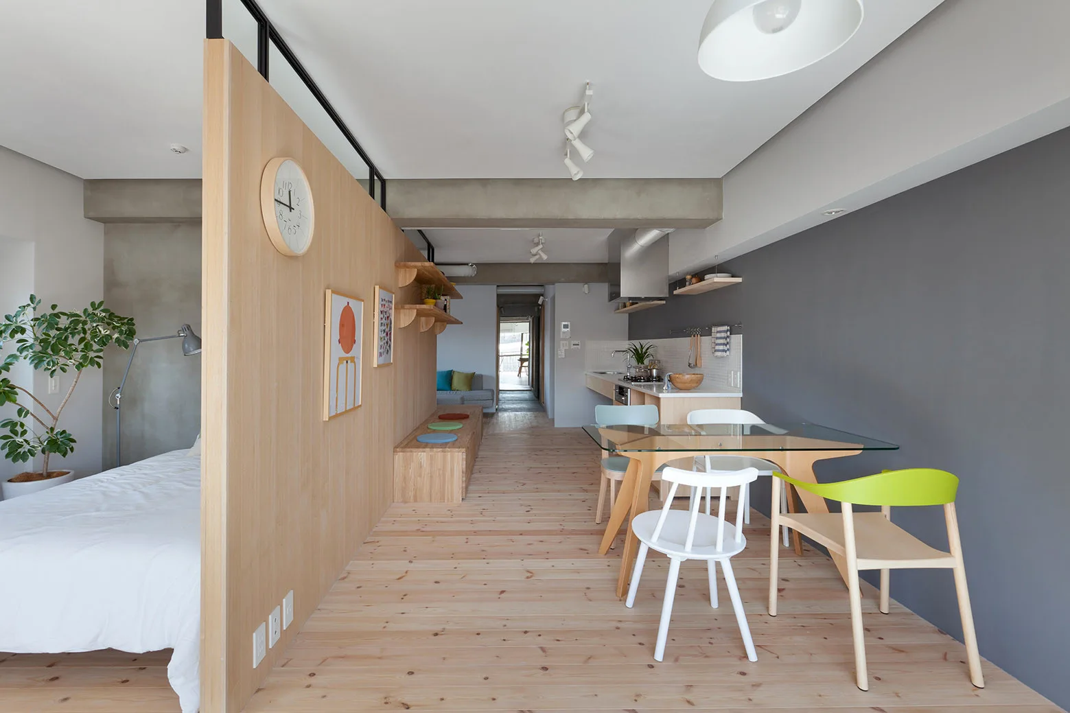 A minimalist studio apartment featuring a light wood partition wall next to a dining area with a glass table and colorful chairs.