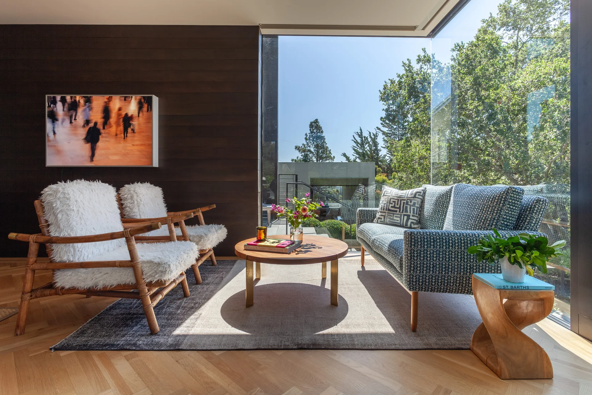 A modern sunlit living room with a blue patterned sofa, two shaggy white armchairs, a wood coffee table, and a large corner window.