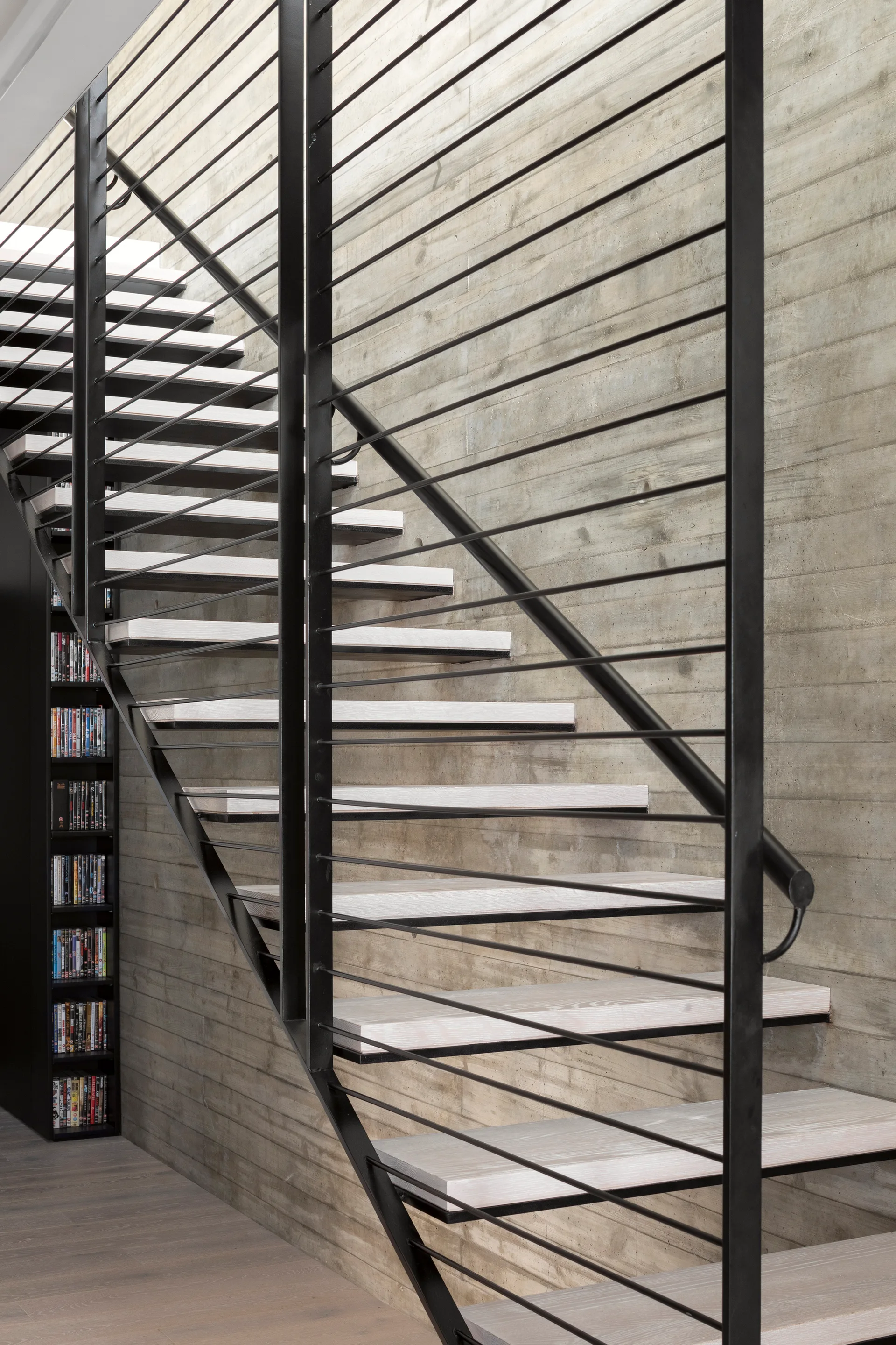 A modern floating staircase with light wood treads and a black metal cable railing stands against a textured, board-formed concrete wall.