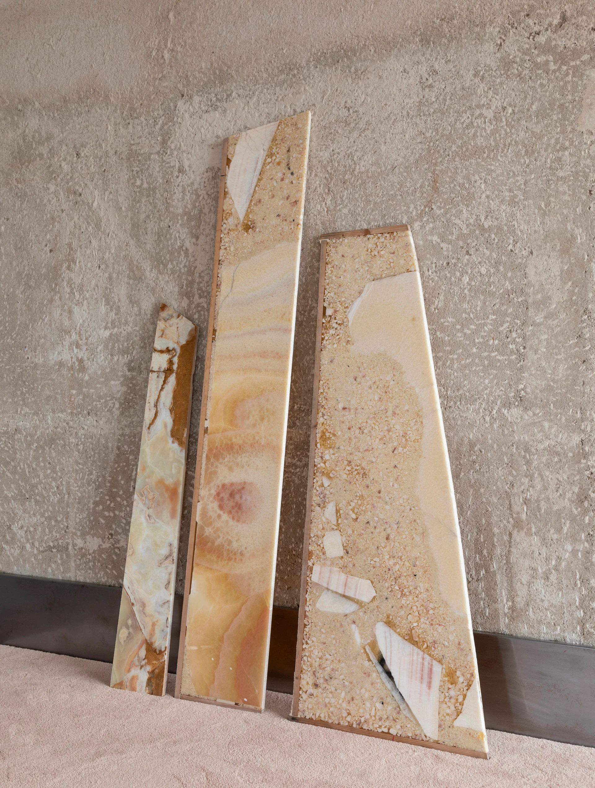 Three tall stone slab samples in warm beige and peach tones lean against a rough, textured plaster wall above a metallic baseboard.