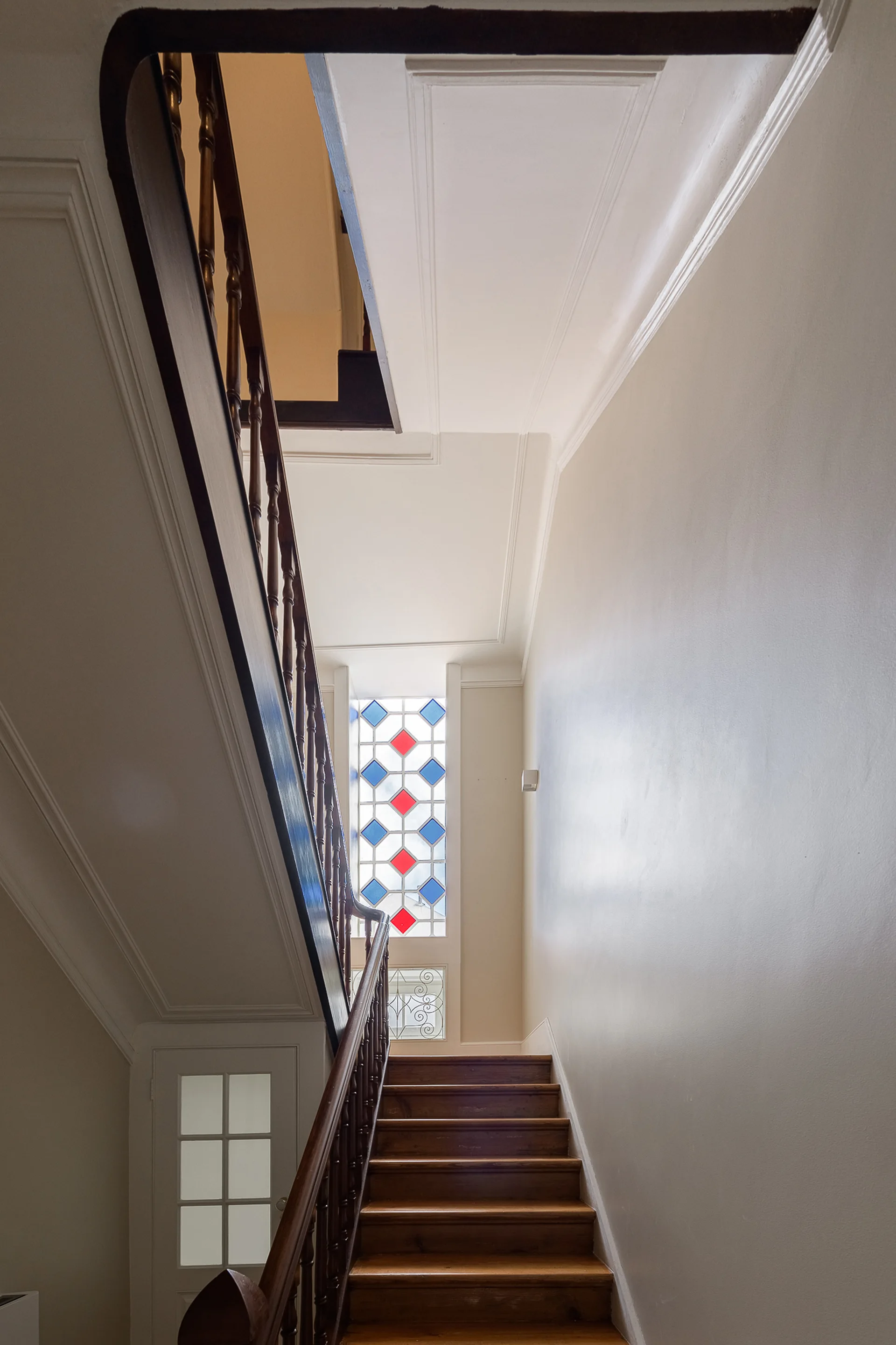 An upward view of a wooden staircase with a dark wood railing, off-white walls with trim, and a colorful geometric stained glass window at the landing.