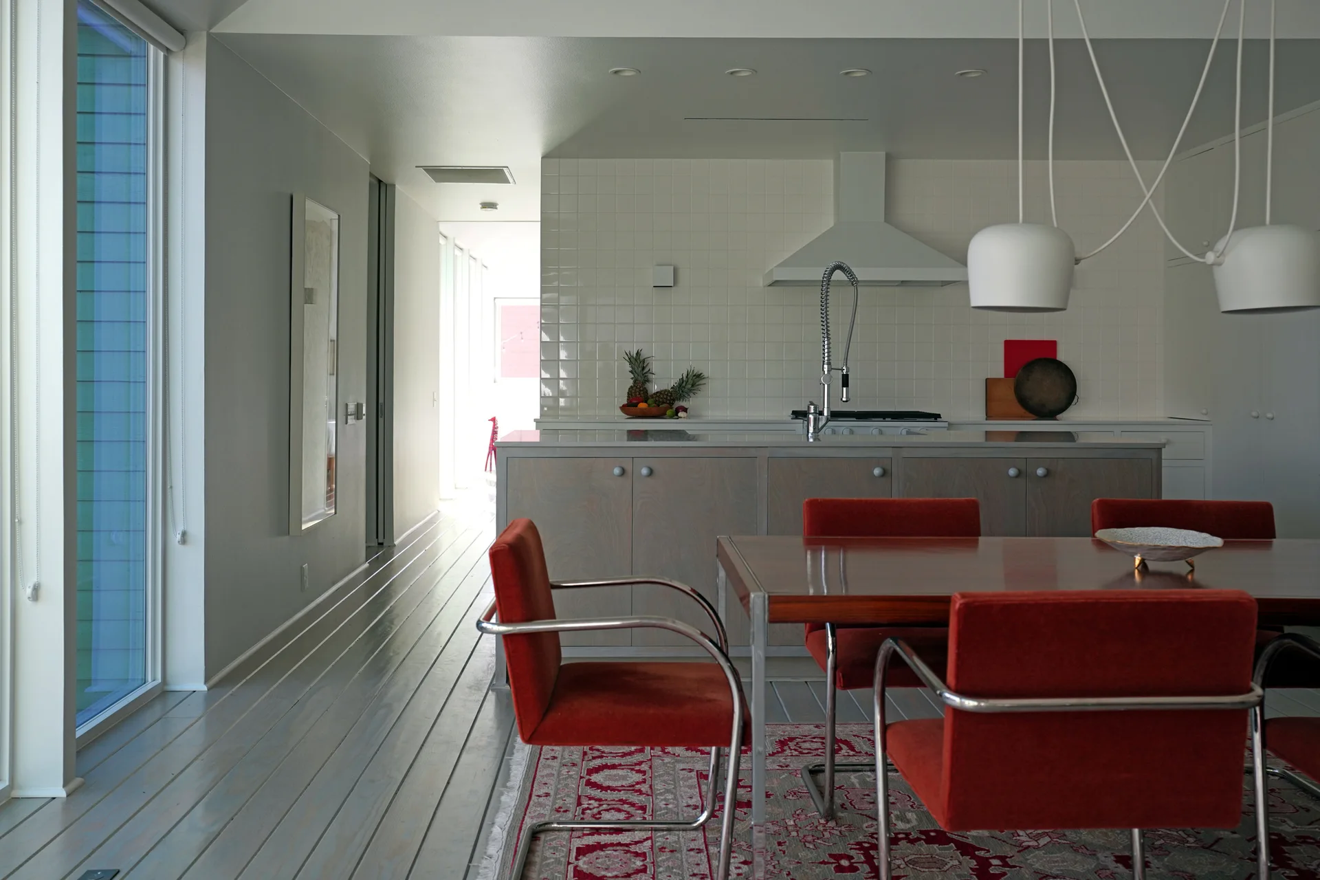 A dining room features red upholstered cantilever chairs around a wood table on a patterned rug next to a white tiled kitchen.